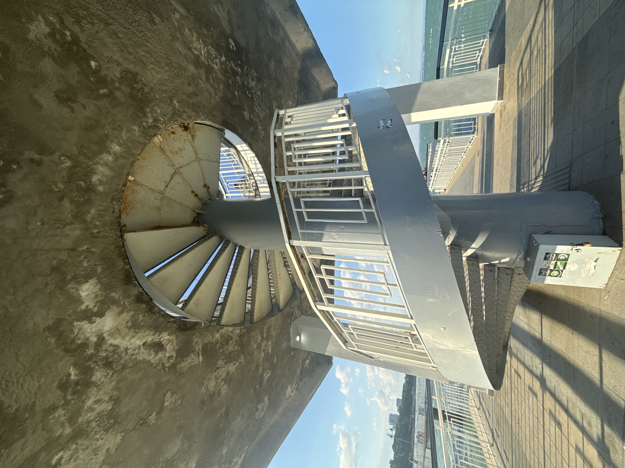 A spiral staircase descends from a circular concrete structure supported by a central column and diagonal pillars. The staircase, painted gray with white railings, has metal treads. The concrete ceiling above shows signs of wear and discoloration. A paved walkway with tiled sections and long shadows stretches alongside the sea, bordered by a white railing. In the distance, a cityscape with buildings and trees is visible on the left, and the sea extends to the horizon under a bright blue sky with scattered clouds. A metal electrical box with stickers is positioned on the walkway near the base of the staircase. The image is taken from a low angle looking up towards the structure. The location is the Sea Garden in Burgas, Bulgaria.