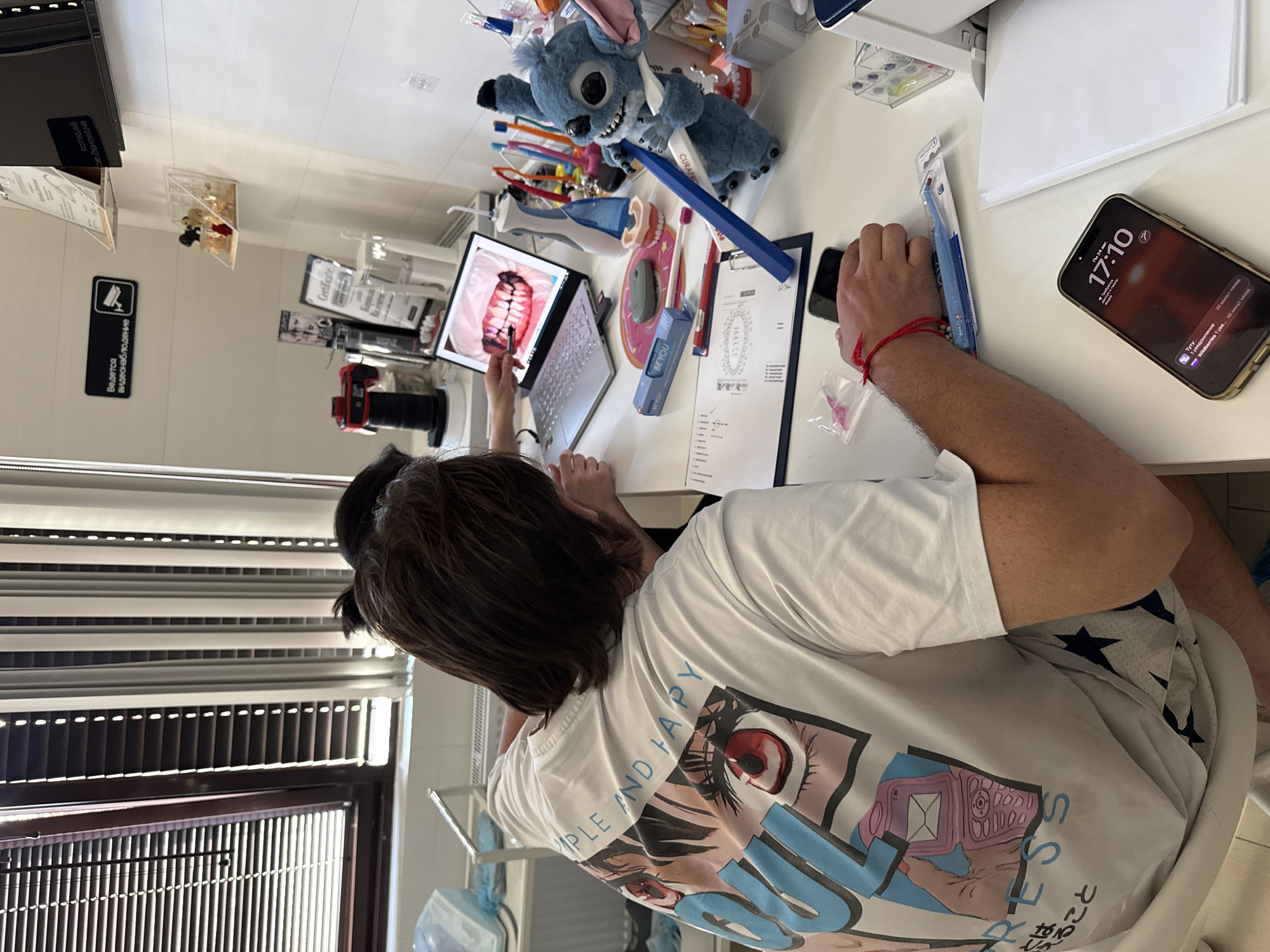 The image depicts an indoor scene, likely a dental office, with two individuals present. The primary focus is on a man from the back, wearing a white t-shirt with an anime-style graphic on the back. He is seated at a desk and appears to be interacting with a laptop which displays an image of teeth. Another person's hand is visible reaching towards the laptop screen, possibly pointing at the display.
The desk is cluttered with various items, including dental hygiene products such as toothbrushes and what appears to be a dental floss dispenser. A stuffed blue character, resembling Stitch from Disney's Lilo & Stitch, is positioned on the desk, holding a blue toothbrush. Also on the desk is a smartphone displaying the time as 17:10. A red string bracelet is visible on the man's left wrist.
In the background, there are windows with blinds, medical equipment, and shelving with items that are not clearly identifiable. A sign with Russian text and a surveillance camera icon is mounted on the wall, indicating the presence of security monitoring. There is no visible indication of the specific location being the city of Mirnoye, Ukraine.