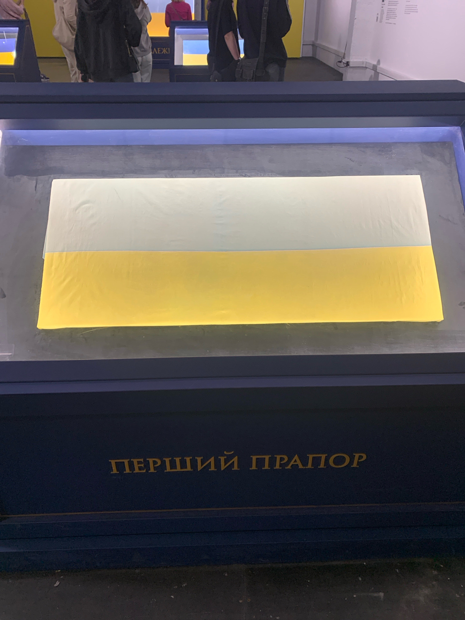 The image shows a display case in a museum, likely in Kyiv, Ukraine, containing a Ukrainian flag with a light blue top stripe and a yellow bottom stripe. The flag is illuminated from below, causing it to glow. The text "ПЕРШИЙ ПРАПОР" (meaning "First Flag") is inscribed in gold lettering on the dark blue base of the display. In the background, several people are visible, appearing to be museum visitors. The environment suggests an indoor exhibition space with art or historical artifacts on display.