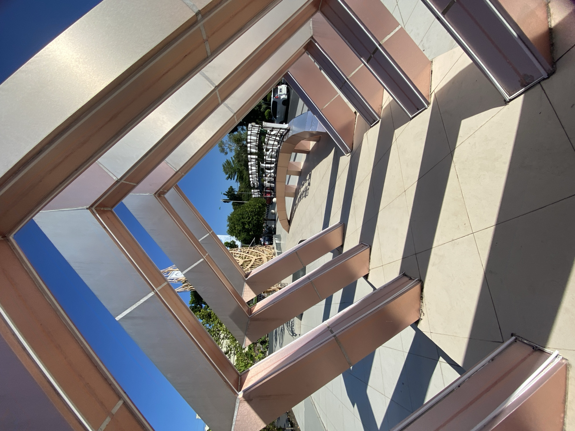 The image shows a modern architectural structure in Banjarbaru, Indonesia. The structure appears to be a sculpture or a gateway, made of metal with a copper-like finish and silver accents. It forms an angular, tunnel-like shape, with its angled beams casting long shadows on the tiled ground. Through the opening of the structure, a street scene is visible. In the background, there is a sign that appears to spell out "BANJARBARU" in large white letters, supported by a curved, multi-tiered structure. Trees line the street, and a white car is parked to the right of the sign. The sky is a clear, bright blue. The overall impression is one of contemporary urban design and public art.
