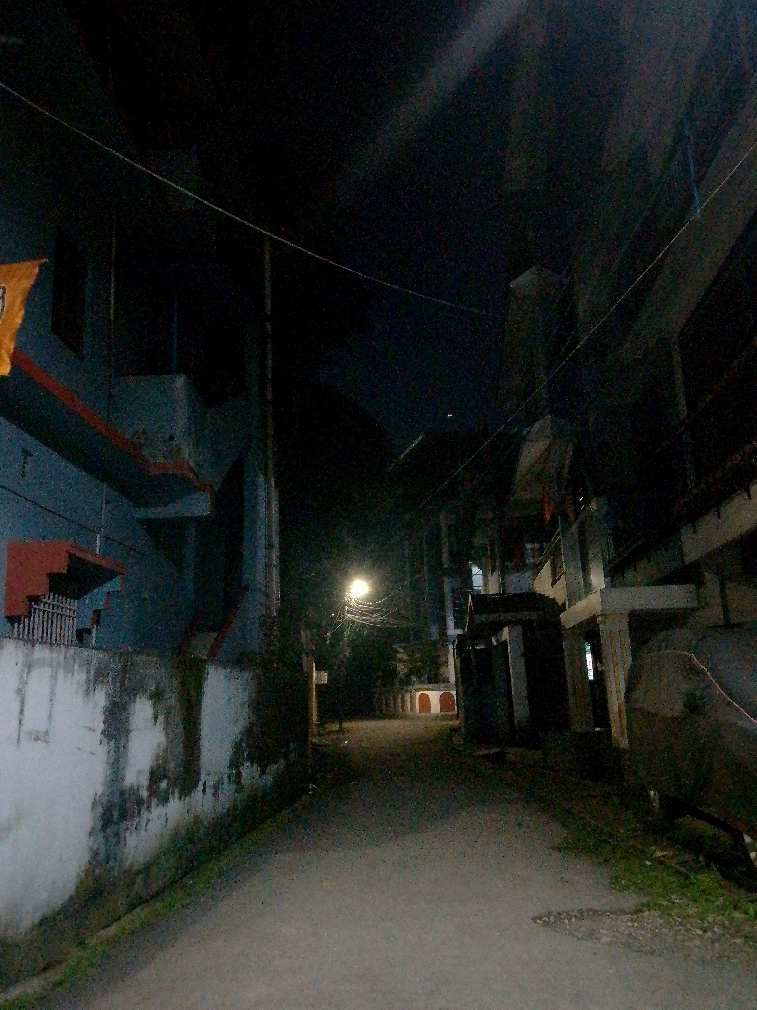 A narrow, unpaved street in Siliguri, India, is illuminated by a single streetlamp. The street is flanked by buildings on both sides. The building on the left is a multi-story structure with a blue facade, a red trim, and a white wall at its base. A portion of an orange flag or banner is visible on the upper left of this building. The building on the right is also multi-story, with a darker, less distinct facade, and appears to have several balconies. The street itself is a dirt or gravel path, with some grass and vegetation growing along the edges. The sky is dark, indicating it is nighttime. There are no people or vehicles visible in the image.
