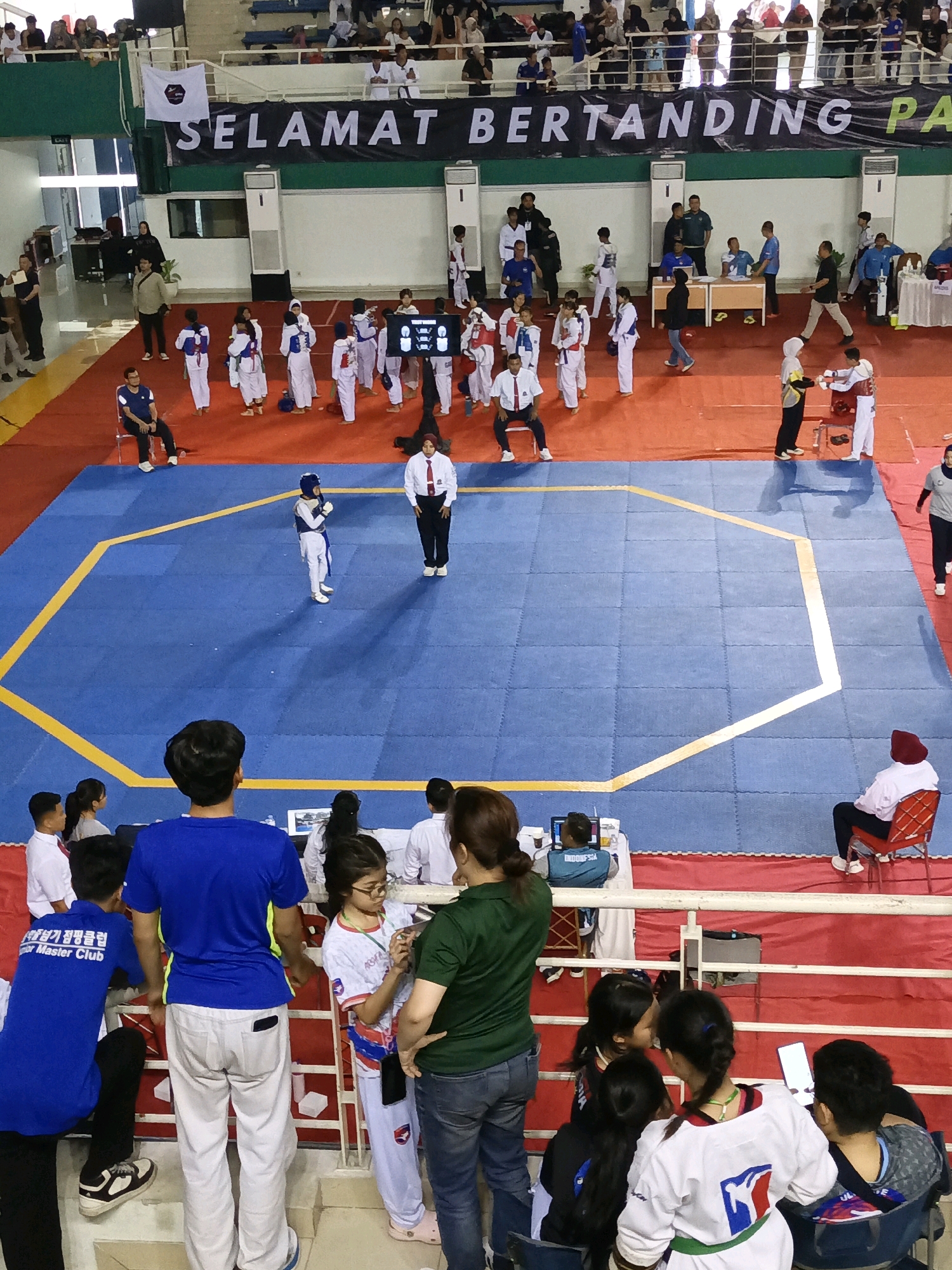 The image depicts an indoor martial arts competition, likely Taekwondo, taking place within a large gymnasium. A prominent banner across the upper section reads "SELAMAT BERTANDING PA" (Welcome to Compete), suggesting the event is in Indonesia. The competition area features a bright blue mat surrounded by yellow boundary lines, set against a red floor. Several individuals are present: athletes in uniform, officials, and spectators.
In the foreground, a group of people are gathered. A young male in a blue shirt and white pants, possibly a coach or organizer, stands with his back to the camera. To his left, a young female in a white uniform with red and blue accents is looking at a phone. Further to the left, another young male in a blue shirt with text on the back is sitting. In the middle ground, several young athletes in white uniforms are lined up, some holding awards or participating in a ceremony. On the competition mat, two individuals in Taekwondo uniforms are engaged in a match, with a referee or official observing. Spectators are visible in the background, seated in tiered seating or standing along the perimeter. The overall environment suggests a formal sporting event with a focus on young participants.
The provided crop images help to identify specific details. One crop shows a closer view of the banner, confirming it is in Indonesia. Another crop highlights a referee in a white uniform with a red tie, standing near the competition mat. Other crops focus on groups of spectators and participants, revealing details like the athletic attire and the presence of electronic scoring displays. The overall impression is a dynamic and organized sporting event for young martial artists.