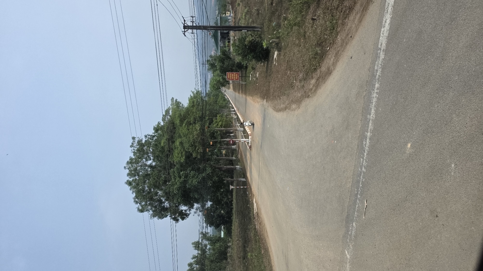 A rural road in Tân Cảnh, Vietnam, stretches into the distance. The road is paved and has a white line marking the edge. Trees line the left side of the road, and power lines run overhead. A warning sign is visible further down the road. The sky is blue. A small, white vehicle is parked on the side of the road in the distance. Vegetation grows along the right side of the road.