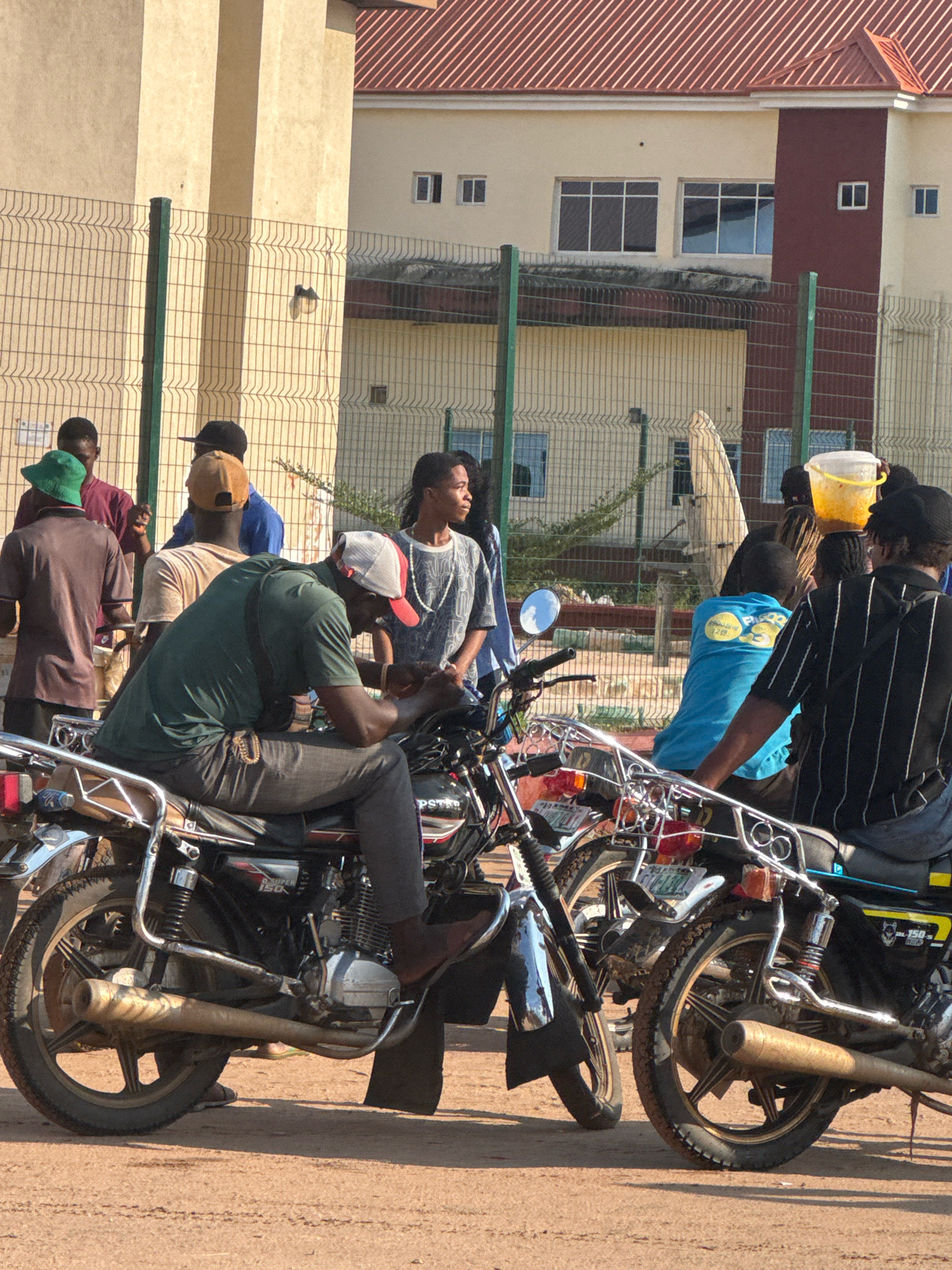 In Ihiagwa, Nigeria, a group of individuals are gathered near motorcycles in front of a beige building with a red-tiled roof. Several men are standing, some in casual attire like t-shirts and caps, while others are seated on motorcycles. One man in a green shirt is seated on a motorcycle in the foreground, looking down at his hands. Another man in a blue shirt is also seated on a motorcycle. A woman in a patterned shirt stands looking to the right. In the background, a yellow bucket is held by an unseen person above a motorcycle. The environment appears to be a street or open area with a wire fence behind the people. The scene suggests a casual gathering or waiting area, possibly related to transportation.