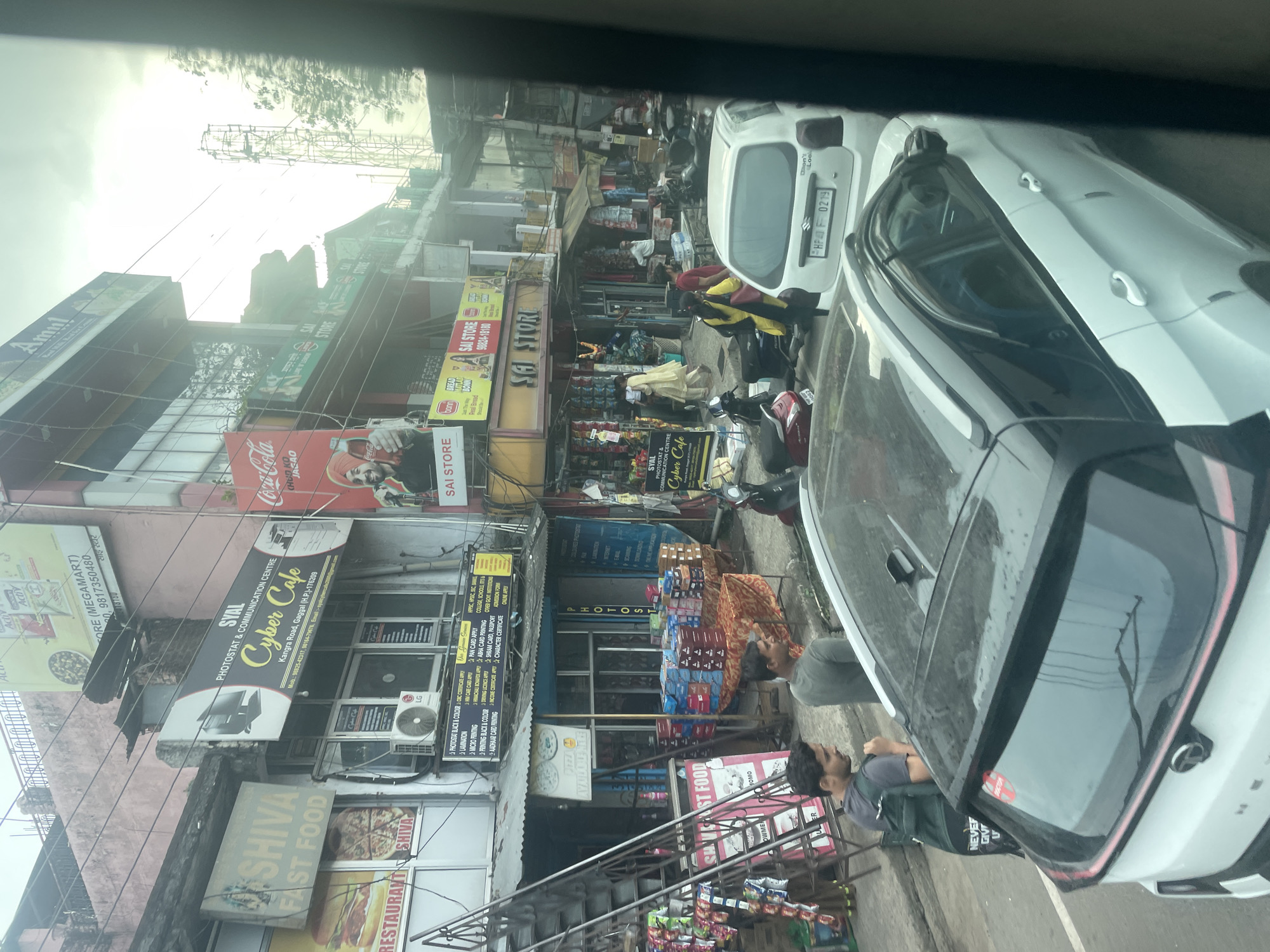 The image captures a street scene in Gaggal, India, likely taken from inside a vehicle. In the foreground, a white car with a black roof is partially visible, with two men standing beside it on the sidewalk. The street is lined with buildings housing various businesses, including a "Cyber Cafe" (SYAL PHOTOSTAT & COMMUNICATION CENTRE) and "SHIVA FAST FOOD". Signage indicates other stores like "SAI STORE" and a Coca-Cola advertisement. Another white car is parked near the "SAI STORE" with the license plate "HP4F 0219". Several motorcycles and scooters are parked along the street. Overhead, a lattice of utility wires crisscrosses the scene. The weather appears overcast.