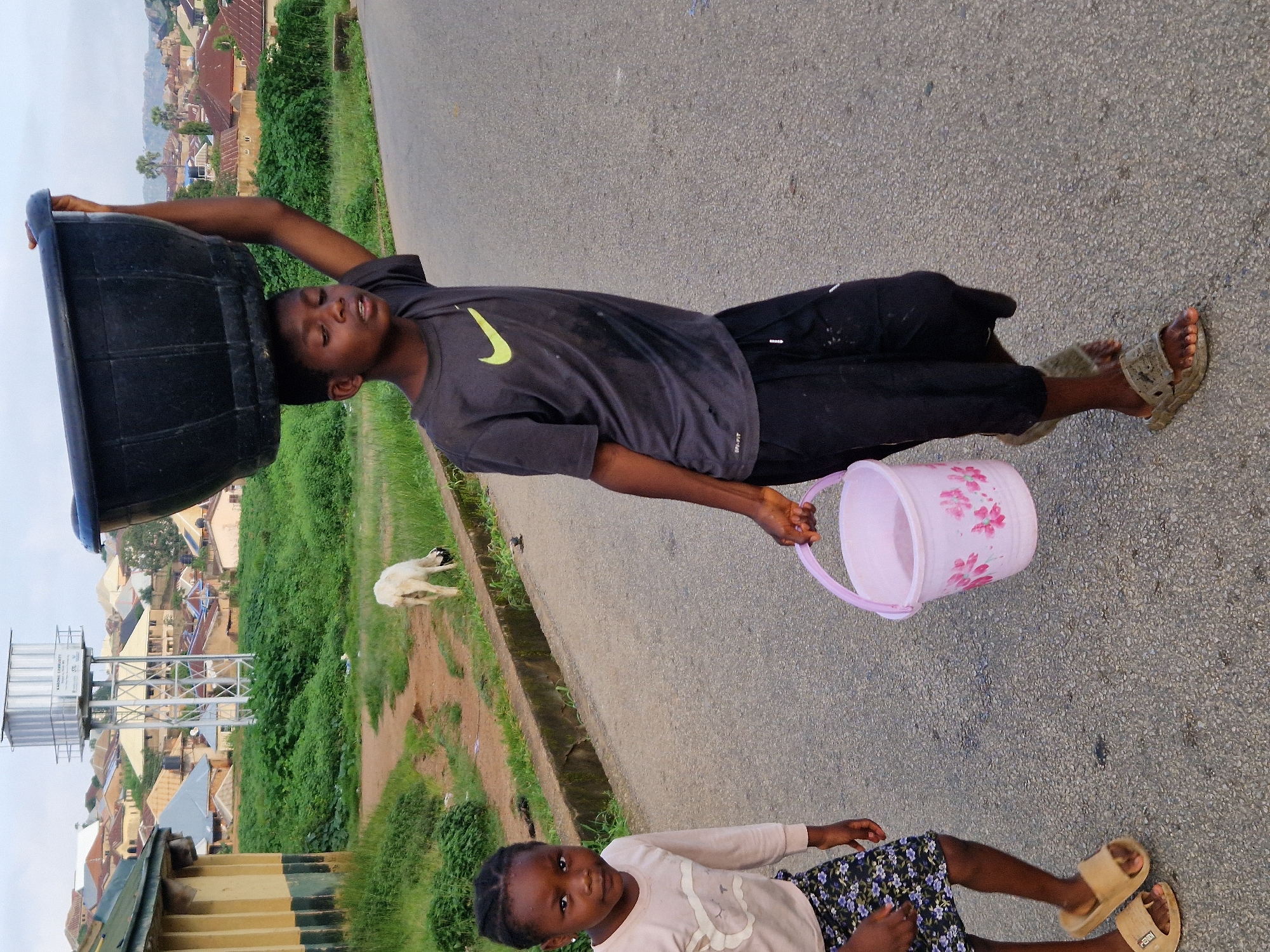 In Kabong, Nigeria, a young boy, estimated to be around ten to twelve years old, walks on a paved road carrying a large black basin on his head. He wears a dark grey t-shirt with a yellow Nike logo and black shorts, and olive-colored sandals. Beside him and slightly behind, a young girl, appearing to be around six to eight years old, walks and looks at the camera. She is wearing a light pink long-sleeved shirt with a graphic of a teddy bear and a dark floral-patterned skirt, along with beige slide sandals. The girl is holding a light pink bucket with a floral design. In the background, a white goat grazes in a grassy area. Beyond the grassy area are buildings with corrugated metal roofs, and a water tower. The sky is overcast.