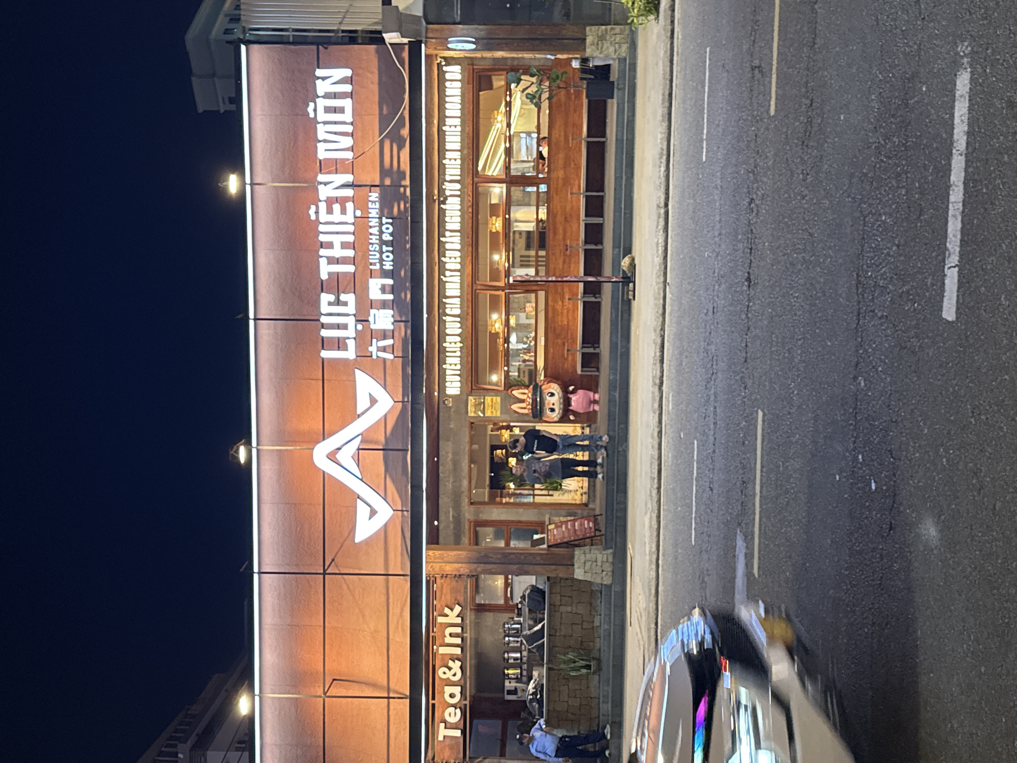 This image depicts the facade of a restaurant and cafe in Đà Nẵng, Vietnam, called "Tea & Ink" and "Lục Thiện Môn," (Liushanmen Hot Pot). The restaurant's sign is in Vietnamese and Mandarin Chinese. Several people are standing near the entrance. A car is blurred in motion on the street.