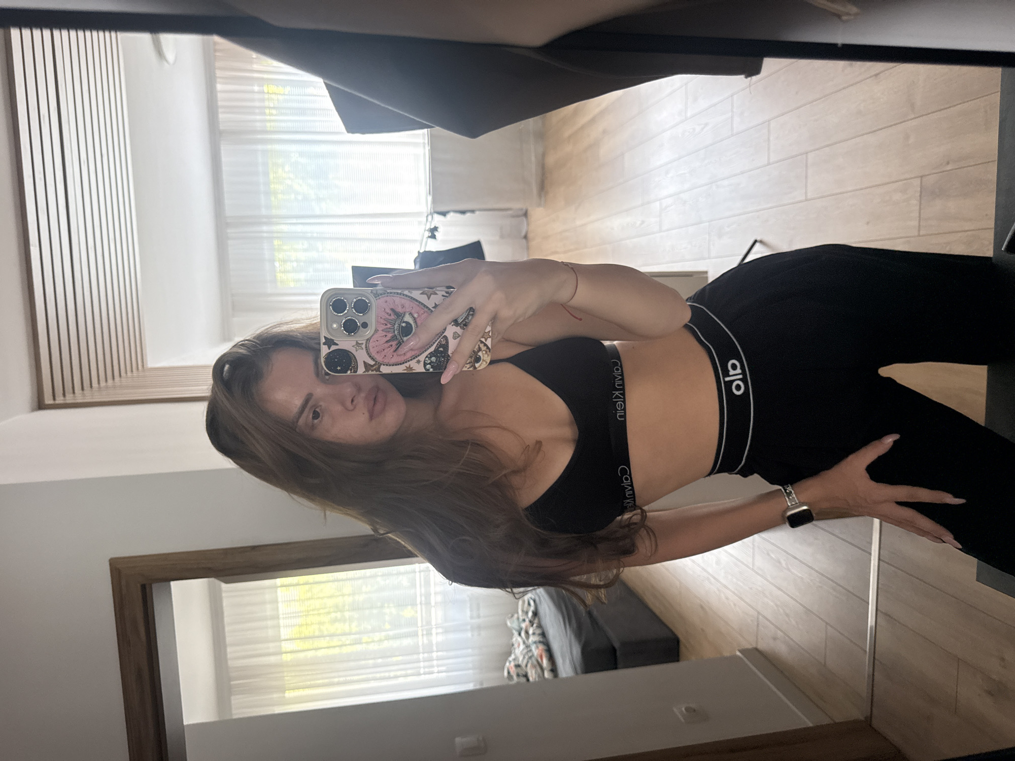This media file shows a young woman taking a mirror selfie in what appears to be a bedroom or dressing area. She is wearing a black sports bra with a white logo and black leggings. Her hair is long and brown, and she is wearing a watch on her left wrist. The background includes a wooden-paneled wall, a large mirror reflecting the room, and what looks like a window with blinds. The overall lighting suggests it is daytime. The setting appears to be a modern, well-lit interior. There is some visible text on the sports bra, which appears to be "CALVIN KLEIN". The woman's face is partially visible, and she has a neutral expression.