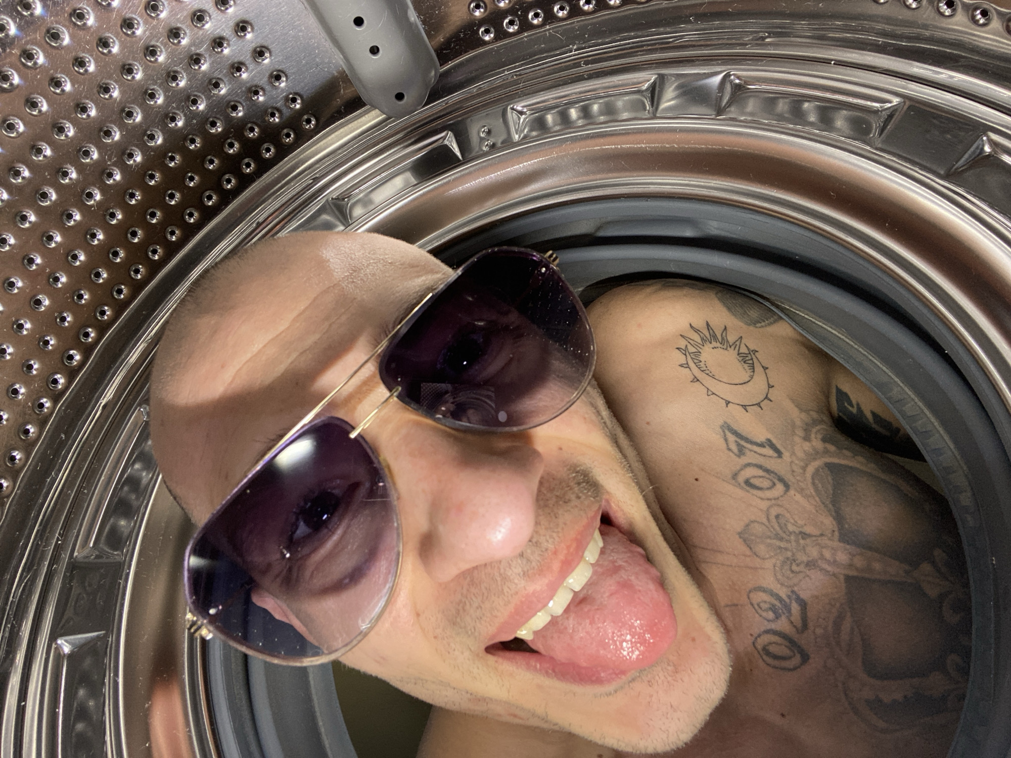 A close-up, distorted view inside a stainless steel washing machine drum features a bald man wearing sunglasses. The man is sticking out his tongue and his head is tilted to the side. He has tattoos on his upper torso, including what appears to be a sun and lettering that reads "2020." The environment is the interior of a washing machine. There is no indication or evidence to establish that the photo was taken in the city of Pleven, Bulgaria.