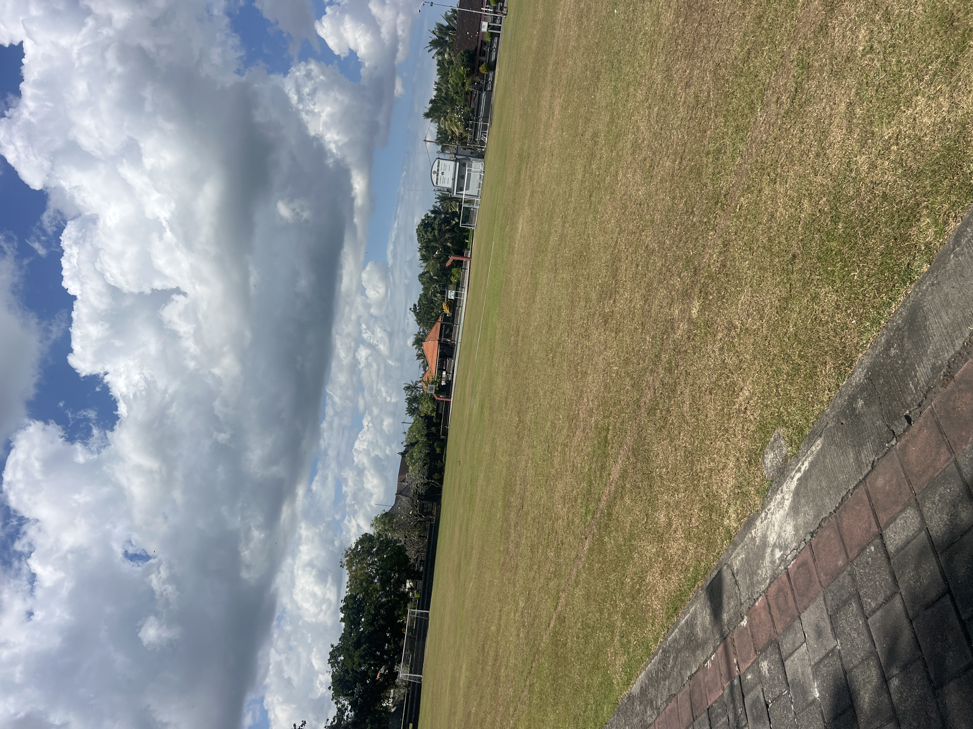 The image shows a wide, dry, grassy field with a paved walkway in the foreground. The field appears to be a sports field, possibly a soccer field, with goalposts visible in the distance. Trees and buildings, including what looks like a small grandstand, are present in the background. The sky is partly cloudy with a mix of bright white cumulus clouds and darker, grayer clouds against a blue sky. The scene suggests a location in Kediri, Indonesia, likely an outdoor recreational or educational facility. There are no people or animals visible in the image. The lighting indicates it is daytime, likely mid-morning or afternoon.