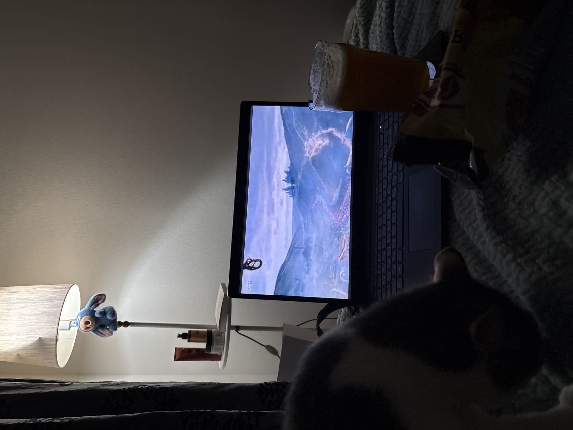 The image shows a scene likely taking place indoors, possibly in a bedroom. A laptop displaying a video game is in the center of the frame, with a glass of beer and a bag of chips resting on it. A lamp with a stuffed animal attached to the side is located to the left. In the foreground, a partially visible cat sits on the bed. The location of Ust-Ilimsk, Russia, cannot be determined from the given image.