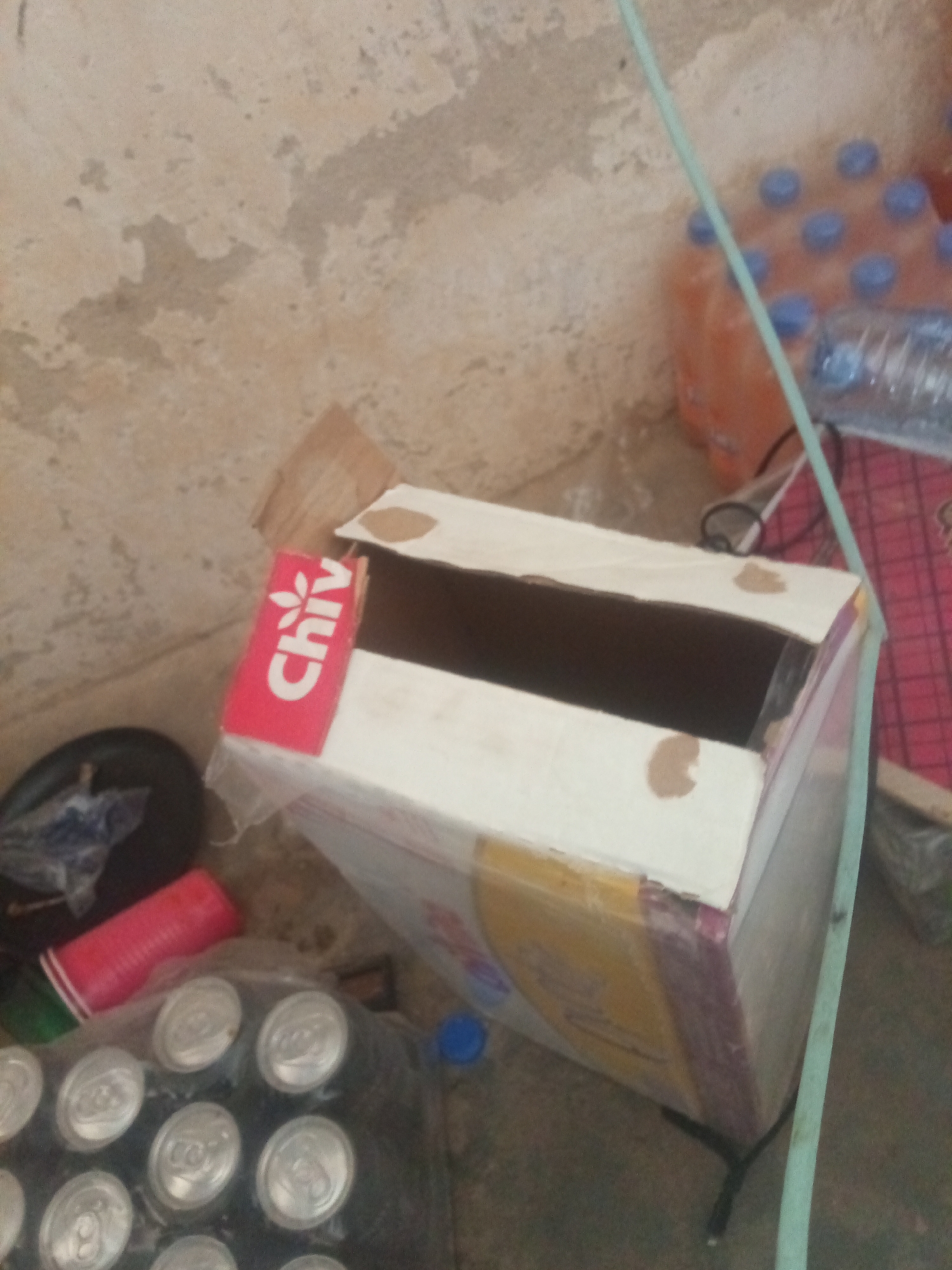 The image depicts an outdoor scene in New Stadium, Nigeria, focusing on a discarded cardboard box with a "Chiv" logo and a pile of canned beverages. The cardboard box, partially open, is positioned in the foreground, showing signs of wear and tear. A collection of canned drinks is visible in plastic wrap beneath the box. In the background, several plastic bottles of what appear to be beverages are stacked, some with blue caps. The environment suggests an area with a weathered concrete or stucco wall and a dirt or concrete floor. A light green hose or cable runs diagonally across the image. Other objects present include a red plastic cup and a black disc-shaped item. The lighting indicates it is daytime.