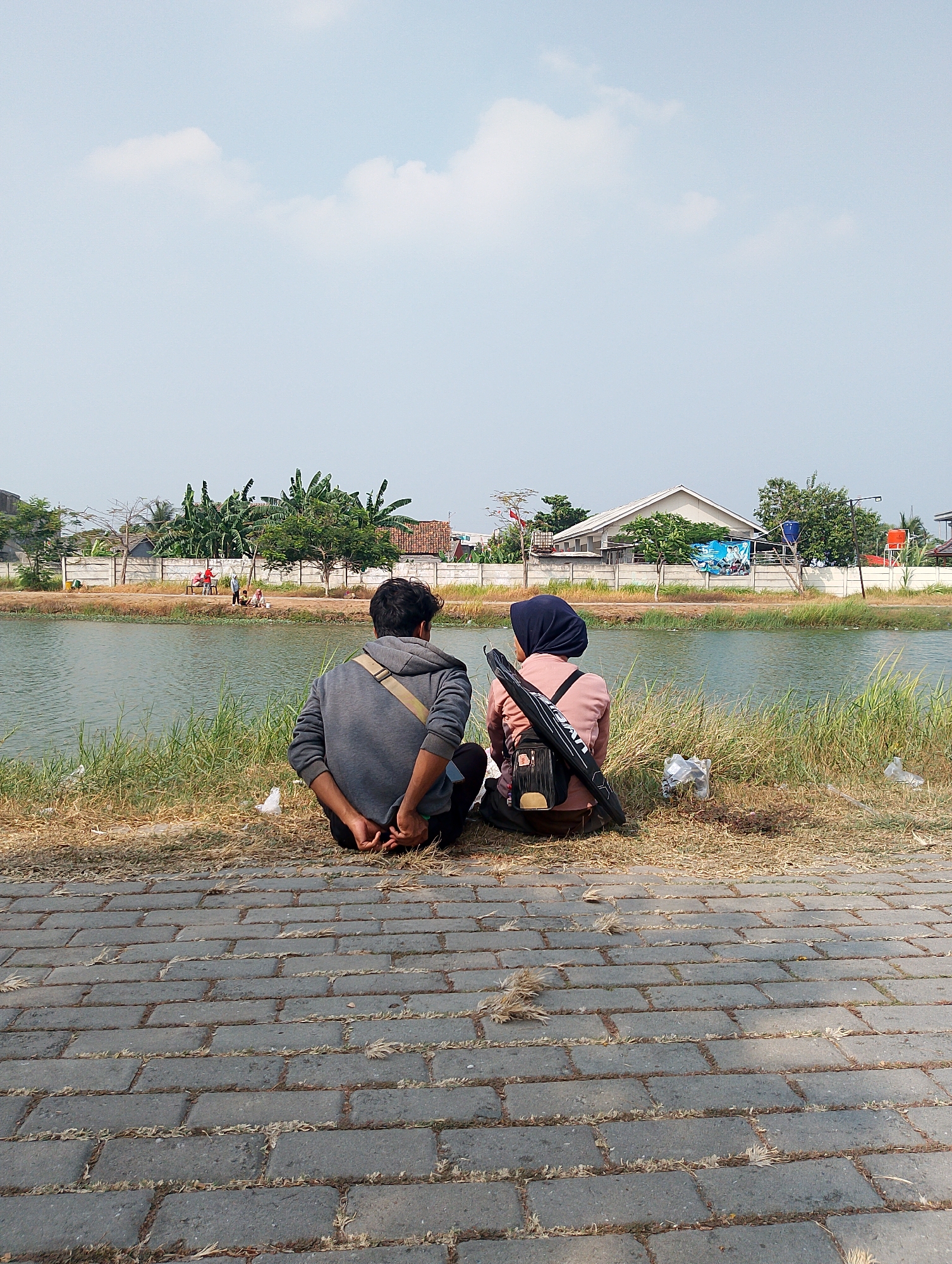 Two individuals, a man and a woman wearing a hijab, sit facing away from the camera on the bank of a body of water, possibly a pond or canal, in Karawang, Indonesia. They are seated on a grassy area with dry grass and some scattered white plastic bags. The man, wearing a gray hooded sweatshirt and dark pants, has his hands clasped behind his back. The woman, wearing a pink long-sleeved shirt and a dark hijab, has a backpack slung over her shoulders and a long black case, possibly for fishing gear or an umbrella, leaning against her. The water reflects the light blue sky, which has scattered white clouds. In the background, across the water, is a fence separating the bank from a row of houses and trees, including banana trees. A few people are visible in the distance on the other side of the water, near the fence. The foreground shows a paved walkway made of grey rectangular stones. The scene appears to be outdoors in a somewhat rural or suburban area during daylight.