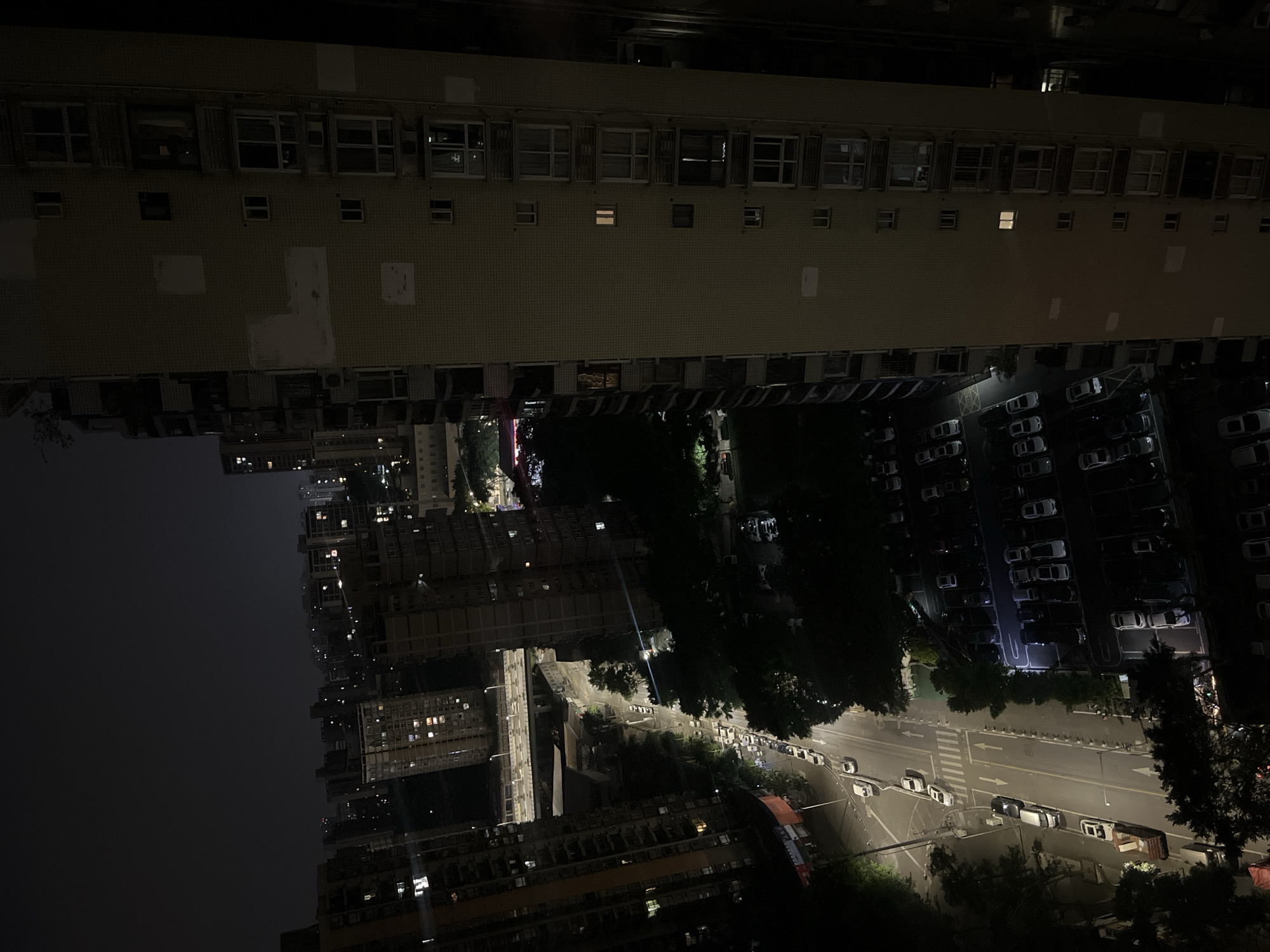 This high-angle, nocturnal photograph captures an urban residential and commercial area in Jiulongpo District, China. The scene is illuminated primarily by artificial lights from buildings, streetlights, and vehicles, against a dark sky.
Dominating the right side of the frame is a tall, light-colored, multi-story building, likely residential, featuring numerous rectangular windows and balconies. Several windows exhibit internal illumination, indicating occupancy. The building's facade is textured with a grid pattern, possibly tiles, and displays patches of lighter coloration or repair.
To the left, a multi-lane asphalt road extends from the lower-left foreground towards the upper-center. White road markings, including a zebra crossing and directional arrows, are visible. Numerous vehicles, predominantly cars, are parked along both sides of the road, and a few are in motion. Overhead streetlights cast bright light onto the road surface and surrounding areas.
Adjacent to the road and below the prominent building on the right, a large outdoor parking lot is densely packed with parked cars, delineated by white parking space lines. This area is also partially illuminated.
In the mid-ground, a multi-level concrete overpass or bridge spans horizontally across the scene, with vehicles discernible on its elevated structure. Beyond the immediate structures, a vast expanse of urban landscape is visible in the background, comprising numerous high-rise buildings with scattered illuminated windows, extending into the darkness. Dark foliage from trees is interspersed throughout the scene, bordering roads and between structures. No individuals are distinctly visible due to the distance and lighting conditions.