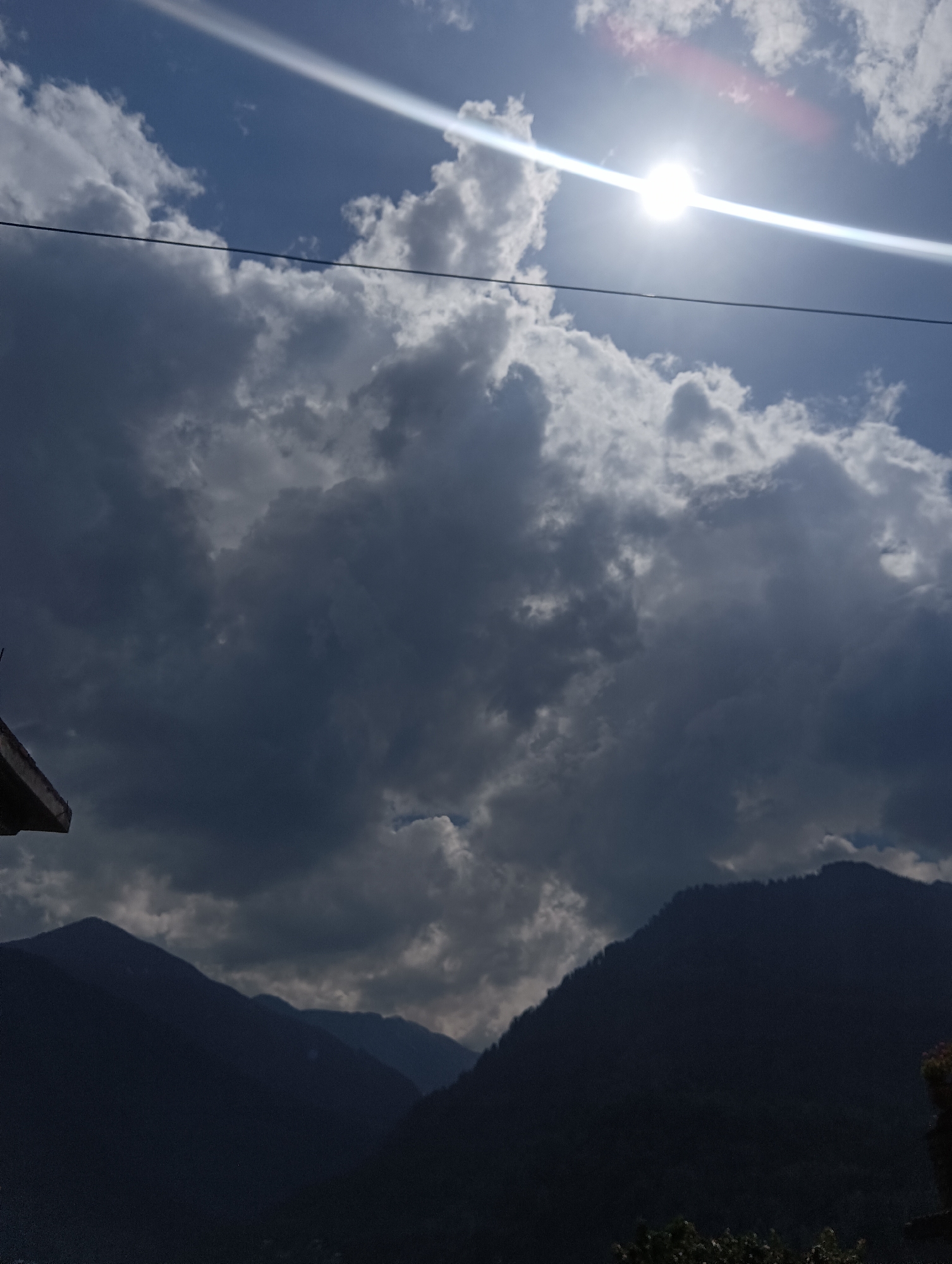 This is a photograph taken outdoors, looking up at a dramatic sky filled with clouds and the sun. The sun is a bright, almost blinding disc positioned in the upper right quadrant, with a strong beam of light cutting diagonally across the frame. Lens flare is visible, adding to the intensity of the sunlight.
The sky transitions from a clear blue in the upper portion to dark, heavy storm clouds that dominate the middle and lower sections of the image. The clouds are varied in texture and shade, with some areas appearing fluffy and white where they catch the sunlight, while others are a deep, ominous grey, suggesting an approaching storm or recent rain.
In the foreground, silhouetted against the sky, are the dark, jagged outlines of mountains. These mountains are covered in a dense forest, though the trees are not clearly discernible due to the low light and distance. The dark shapes of the mountains create a stark contrast with the bright sky. A single black wire runs horizontally across the upper part of the image, possibly a power line or telephone wire, adding a man-made element to the natural landscape. On the far left, a small portion of what appears to be a concrete structure, possibly part of a building or scaffolding, is visible, also silhouetted.
The time of day appears to be late afternoon or early evening, given the position of the sun and the dramatic interplay of light and shadow. The weather is mixed, with bright sunshine and impending or passing stormy weather. The location appears to be mountainous, likely a rural or wilderness area. No people are visible in the image, nor is any text. The overall mood is one of awe and perhaps a touch of foreboding, due to the powerful sun and the dark, heavy clouds.