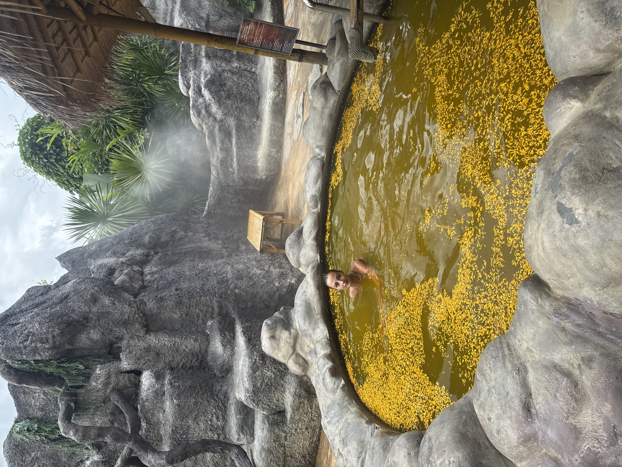 A woman is chest-deep in a round, hot spring pool filled with yellow flower petals, surrounded by artificial rock formations. The pool is located in Ban Khlong Khian, Thailand, based on the location metadata. There is steam rising near a small bamboo chair and thatched shelter. A sign with text is posted nearby.