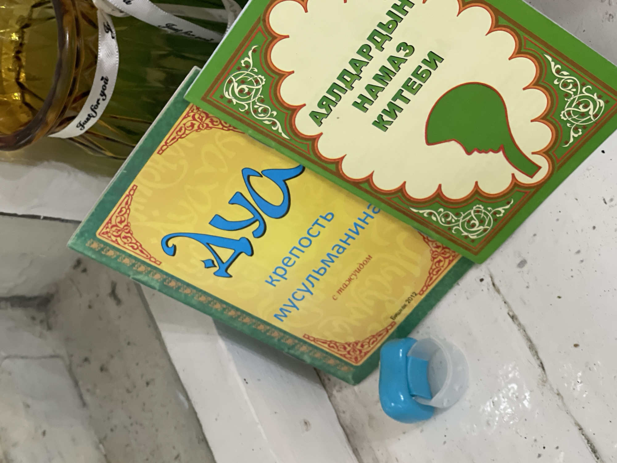 In a close-up, eye-level shot, two books are placed on a white, textured surface that appears to be a windowsill or a similar flat surface. To the left, a small, blue plastic object, possibly a baby pacifier cover, rests on the surface.
The book in the foreground, on the right, has a green cover with an ornate, cream-colored border. The title, in green Cyrillic script, reads "АЯЛДАРДЫН НАМАЗ КИТЕБИ" which translates to "Women's Prayer Book". A stylized green silhouette of a woman's profile is centered on the cover.
Behind it, slightly to the left, is another book with a yellow cover and a green border. The title is in large, blue script, possibly "Дуа" (Dua), followed by smaller blue text that appears to be "крепость мусульманина" (strength of a Muslim) and "с таджуидом" (with Tajwid). At the bottom of this book, a small inscription in Cyrillic appears to read "Бишкек 2012", indicating Bishkek, Kyrgyzstan as the place of publication in 2012.
To the upper right, a portion of a glass vase with an amber tint is visible. A white ribbon is tied around the vase, with the words "Just for you" printed on it in black script. The overall lighting suggests an indoor environment, with soft light falling on the objects.
