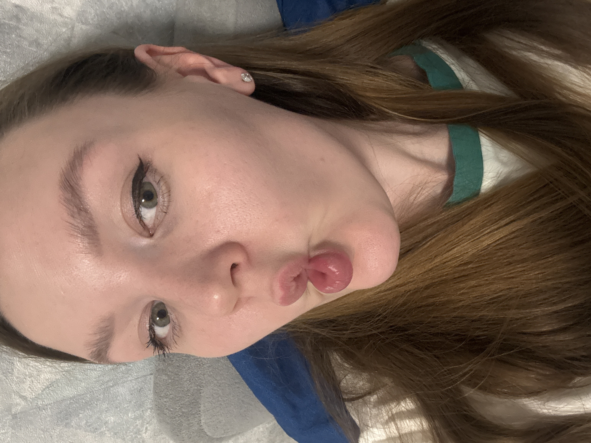 A young woman with light skin and long brown hair is lying on her side, facing the camera, with a pouting expression. She has light green eyes and distinct eyebrows. She is wearing a white shirt with a green collar. Her ear is visible, and she has a stud earring. The background consists of a textured, light-colored surface, possibly a bedsheet or other fabric, with a blue object partially visible beneath her head. The image does not contain any details connecting it to Izhevsk, Russia.