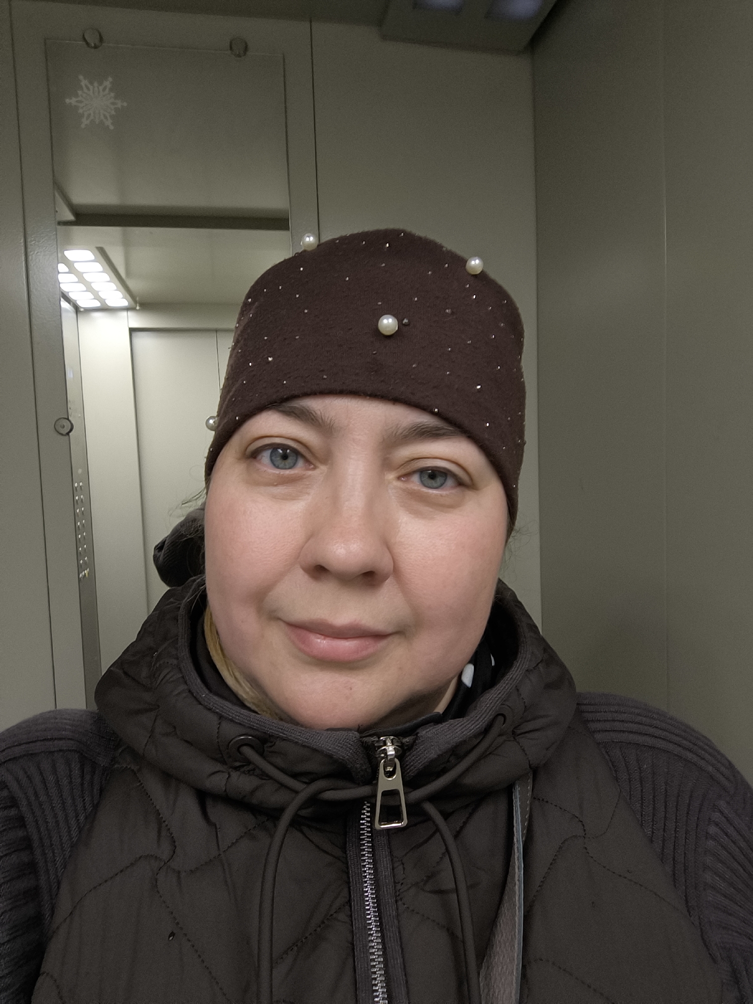 A medium shot photograph shows a woman in Khimki, Russia, based on the details observed in the image. She is centered and faces the camera. The woman has light skin, blue eyes, and a slight smile. She is wearing a dark brown knit hat embellished with white pearls and a brown jacket with a zipper. The background reveals a closed elevator. The elevator's interior is utilitarian, featuring steel panels and a small, rectangular mirror affixed to the left panel, which has a snowflake sticker on it. An array of illuminated buttons is also visible on the left. The lighting is even, with the light source seemingly overhead within the elevator.
