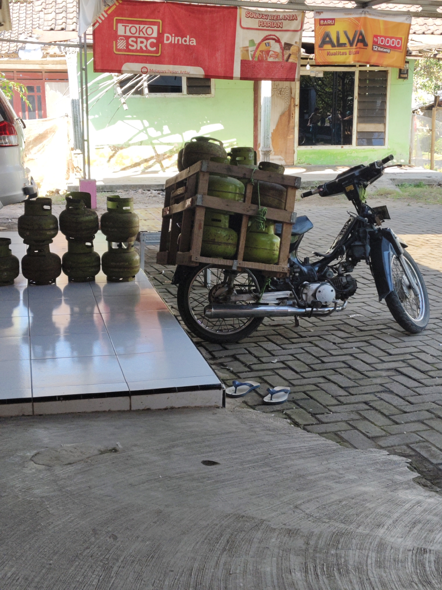 In Bangsalsari, Indonesia, a motorcycle is parked on a brick-paved street in front of a small shop. The motorcycle is carrying a wooden crate filled with several green LPG gas cylinders, stacked three high. In the foreground, on a tiled surface, more LPG cylinders are arranged. A white car is partially visible on the left side of the image. The shop has a light green facade and a red banner hanging above it, with text and logos visible. The scene appears to be outdoors on a sunny day, with shadows cast on the ground.