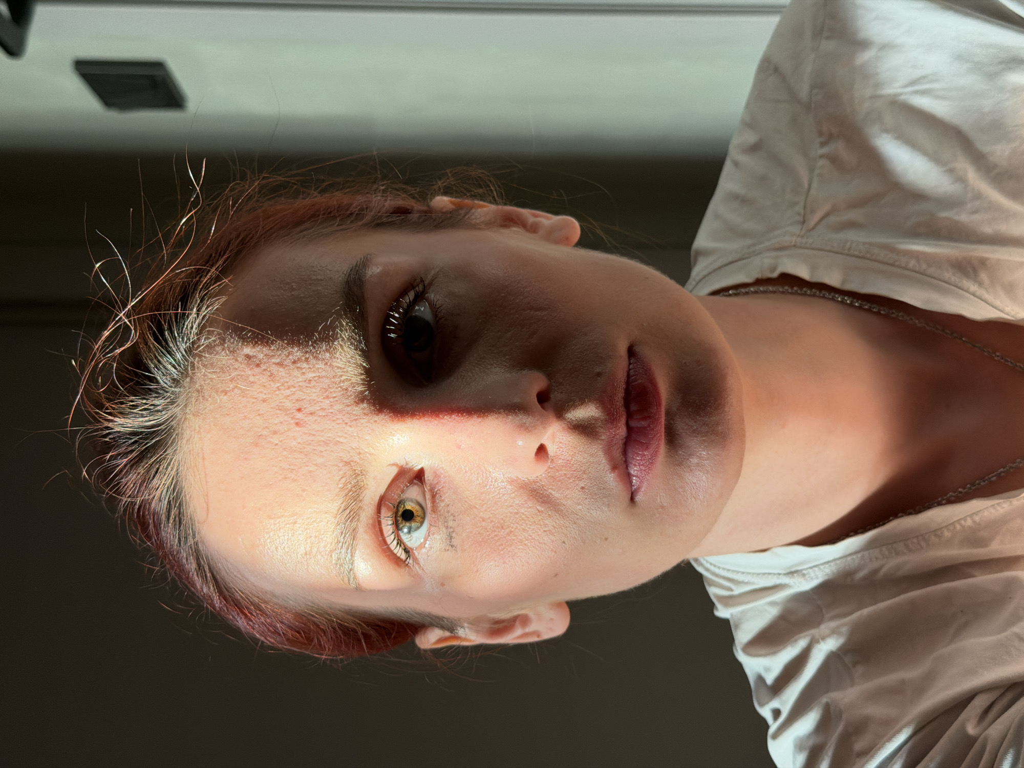 A close-up, low-angle portrait shows a fair-skinned person with red hair and green eyes. The person is wearing a white shirt and a silver chain. The left side of the face is illuminated by bright light, creating a stark contrast with the shadowed right side. The background is dark and out of focus. A square, dark object is visible in the upper left corner of the image. There is no indication that this image was taken in the city of Chaikovsky, Russia.