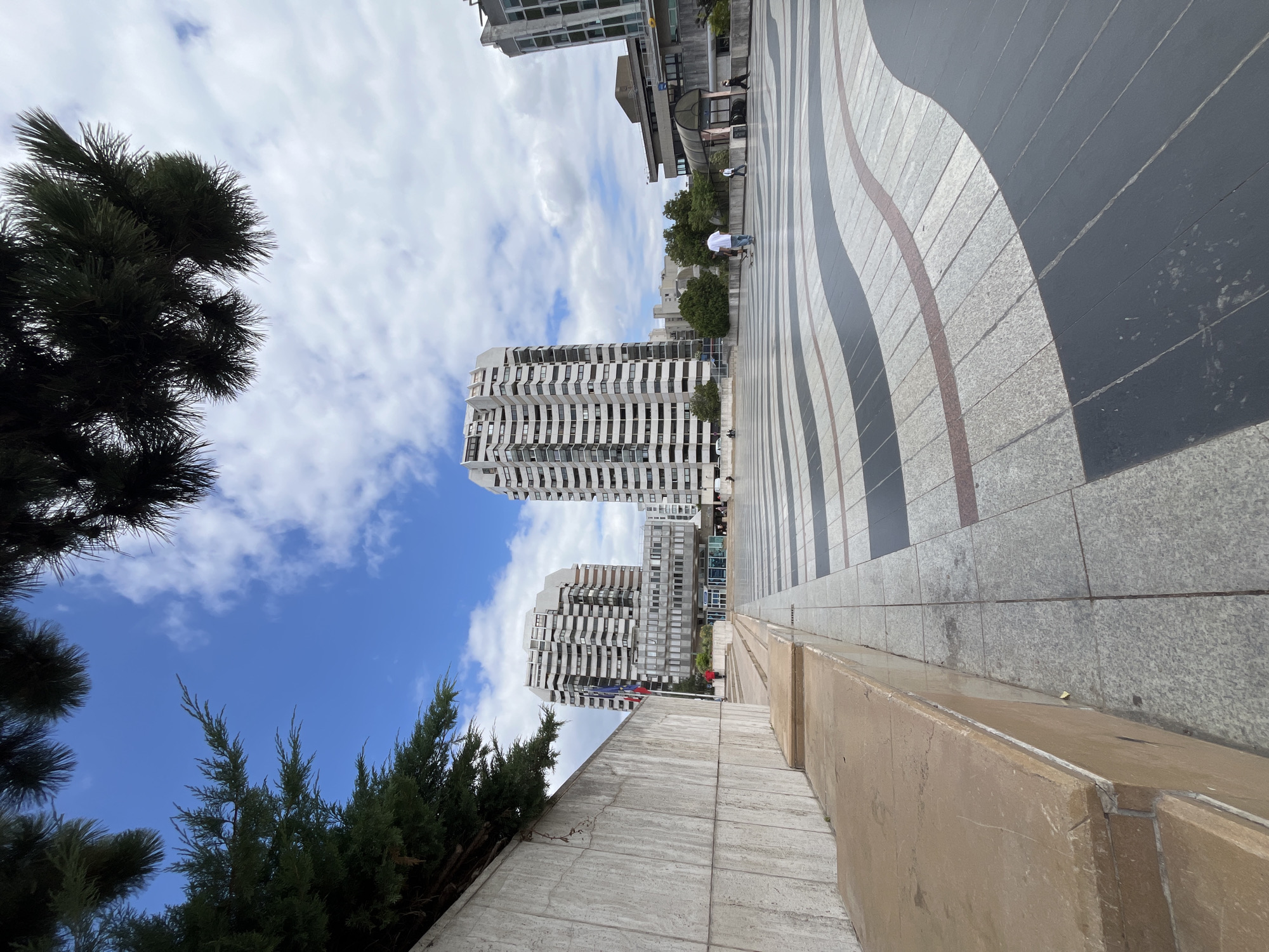 The image depicts a wide, paved plaza with a distinctive wave-like pattern of dark gray and light gray rectangular tiles, interspersed with a thin red tile border. In the background, two tall, modern, multi-story white buildings with numerous windows dominate the skyline. A skybridge connects these two buildings. To the left, a low, textured concrete wall with a raised stone seating area runs along the edge of the plaza. Green trees with dense foliage are visible above the wall and in the middle ground. The sky is partly cloudy with patches of blue.
Several people are present in the plaza. In the mid-ground, a person in a white t-shirt and blue jeans is skateboarding. To the right, two individuals are walking, one wearing a white shirt and the other dark clothing. Further in the distance, more people are scattered throughout the plaza, some walking and others standing. Two flags are visible on flagpoles near the base of the building on the left. The scene appears to be an urban public space, likely in the city of Créteil, France, given the architectural style of the buildings which is characteristic of the area, particularly the "Chaudron" district.