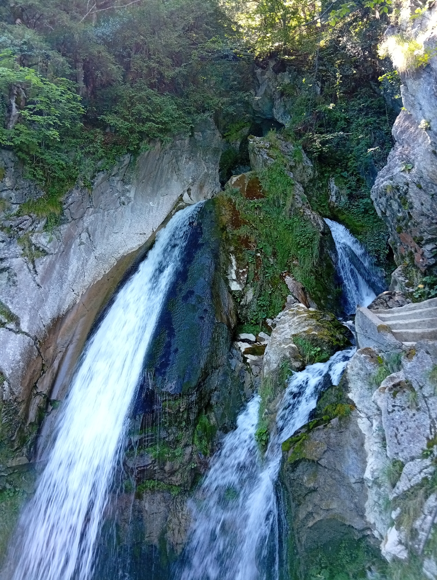 This is a photograph of a cascading waterfall in Emmetten, Switzerland, taken during the daytime. The waterfall flows down a rocky, moss-covered mountainside, surrounded by lush green vegetation. Sunlight filters through the trees at the top of the frame, illuminating parts of the scene. To the right of the waterfall, a set of concrete stairs is visible, leading upwards along the mountainside. The water is a blur of white due to the long exposure, indicating a smooth, continuous flow. The rocks are varied in texture and color, with some appearing smooth and others rough and fractured. The overall impression is one of a natural, serene, and somewhat wild environment.