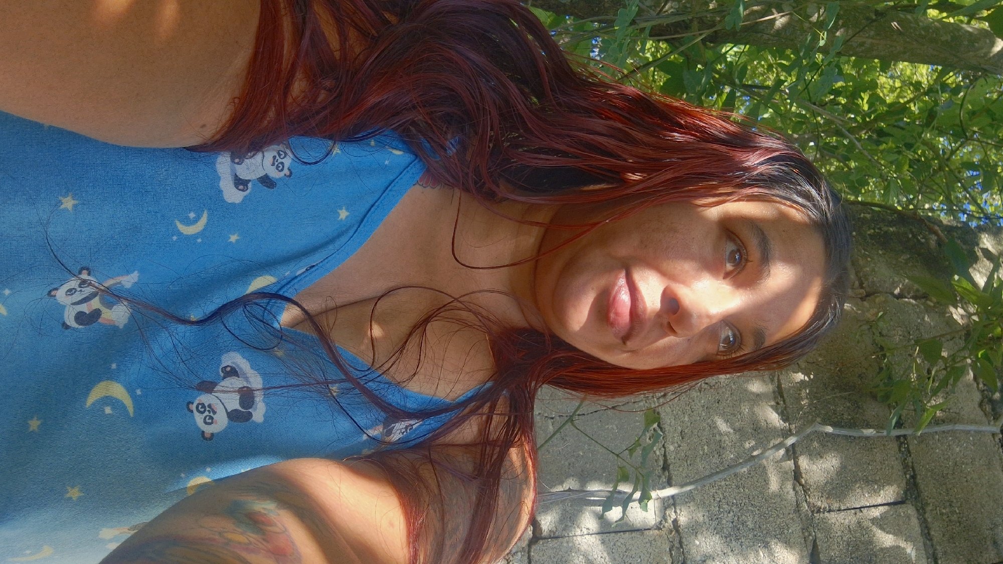 In Vallecito, Venezuela, a woman with long, dark red hair poses outdoors, looking upwards. She is wearing a blue tank top adorned with a pattern of pandas and crescent moons. The background consists of lush green foliage and a textured, light-colored wall, suggesting an outdoor setting. Sunlight casts shadows across the scene, highlighting the woman's face and upper body.