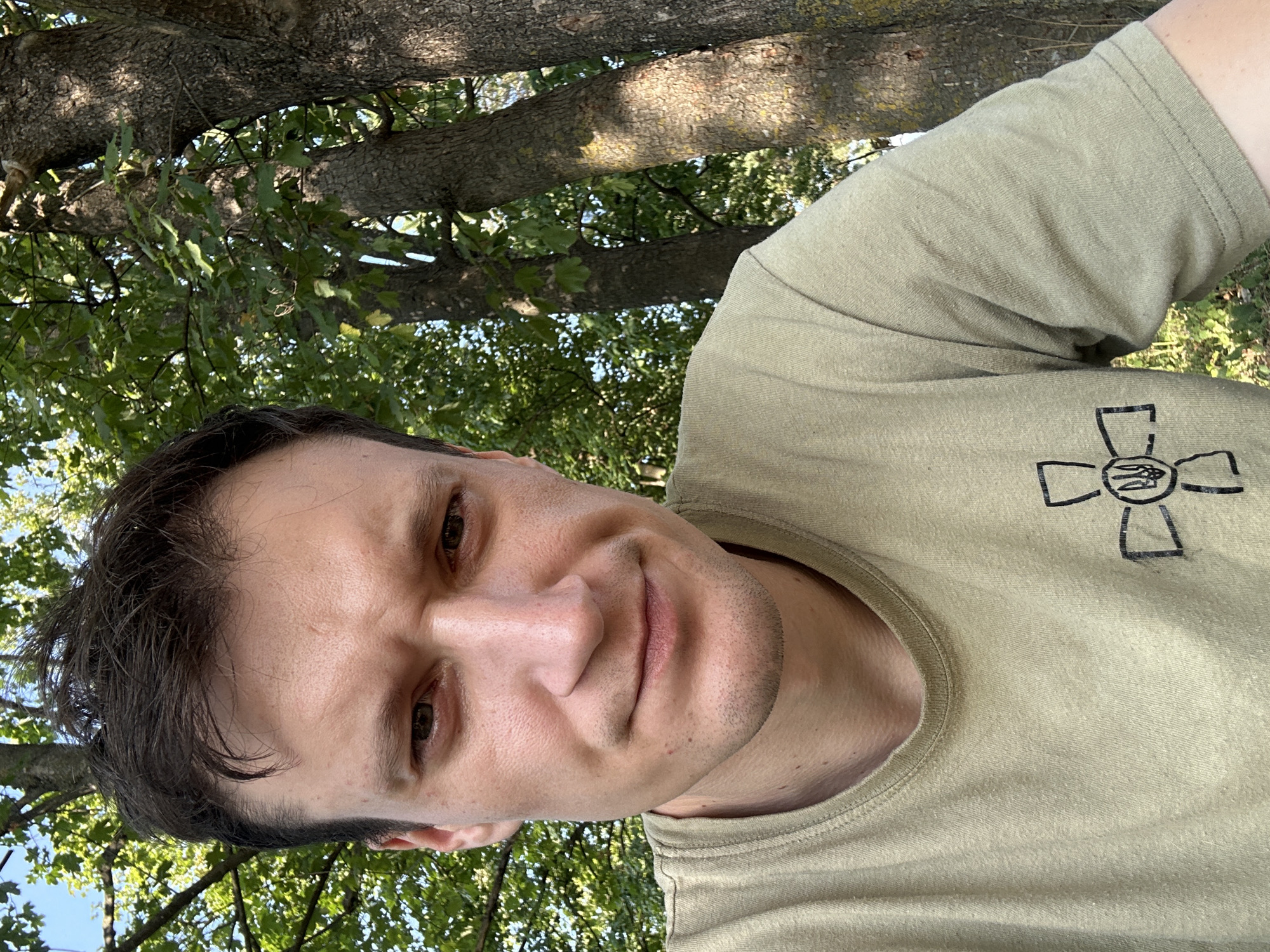 A man is taking a selfie outdoors in a wooded area, likely in Makiv, Ukraine, as indicated by the symbol on his shirt which is the trident, the national emblem of Ukraine. He is wearing a light olive-green t-shirt with the emblem printed on the left side of his chest in black. The man has short, dark brown hair, dark eyes, and is looking directly at the camera with a slight frown. His skin appears fair and slightly tanned, with some texture visible on his forehead and cheeks. He has stubble on his chin and upper lip. The background consists of several large trees with textured bark and lush green foliage. The lighting suggests it is a sunny day, casting shadows from the trees. The ground is covered with grass and some dry leaves.