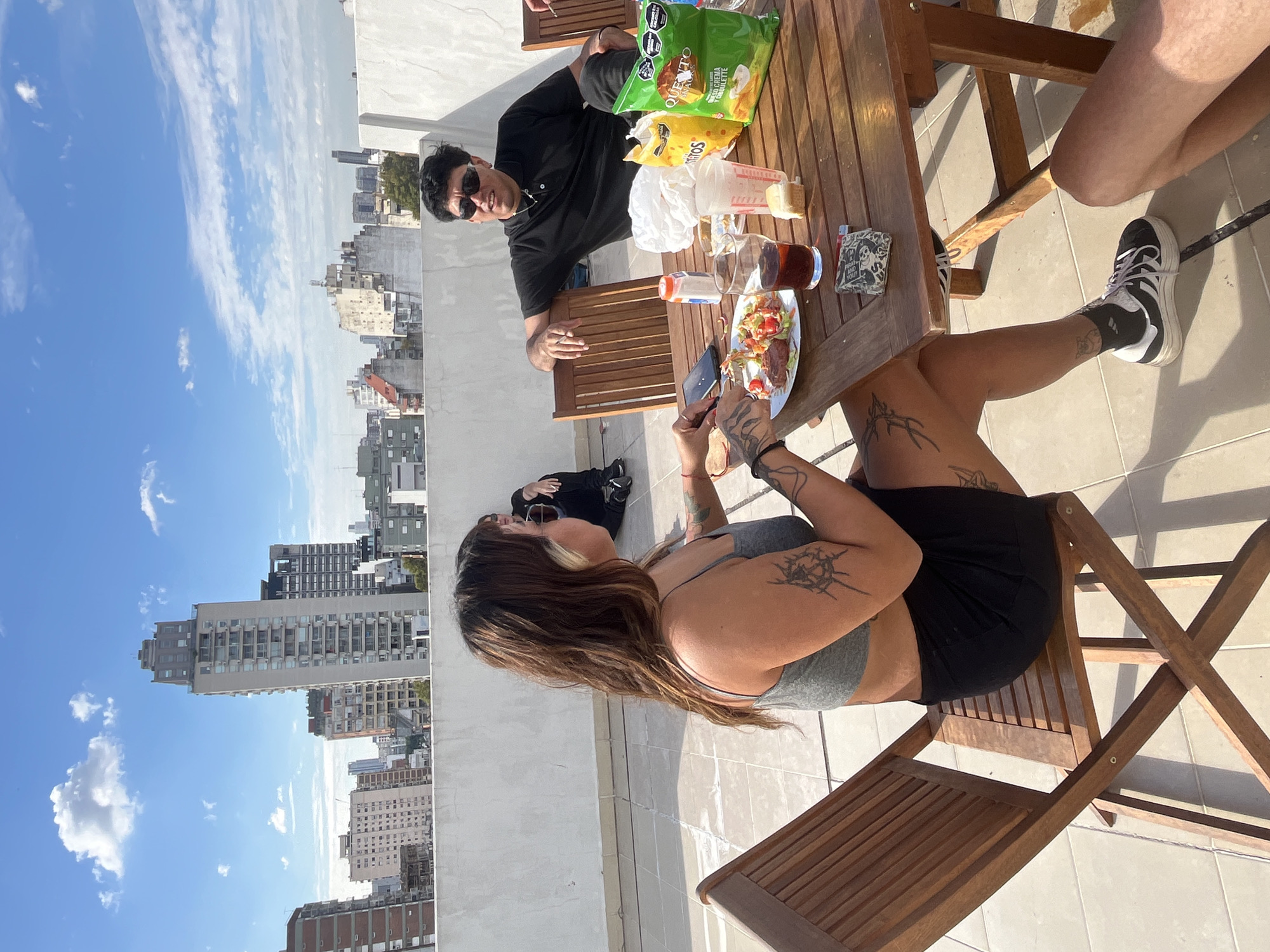 The image depicts an outdoor gathering on a rooftop terrace in Buenos Aires, Argentina, suggested by the city skyline in the background. A wooden picnic table occupies the center of the frame, laden with snacks including bags of chips. Two people are visible around the table: A man wearing sunglasses and a black shirt sits opposite a woman with tattoos on her arms and legs. She is wearing a gray top and black shorts, seated on a wooden chair, and appears to be photographing the food with a mobile phone. The woman's legs are positioned to the side, and her feet are clad in white and black sneakers. Another figure is visible on the ground behind the table, but their features are indistinct. The scene is set against a backdrop of blue sky and cityscape.