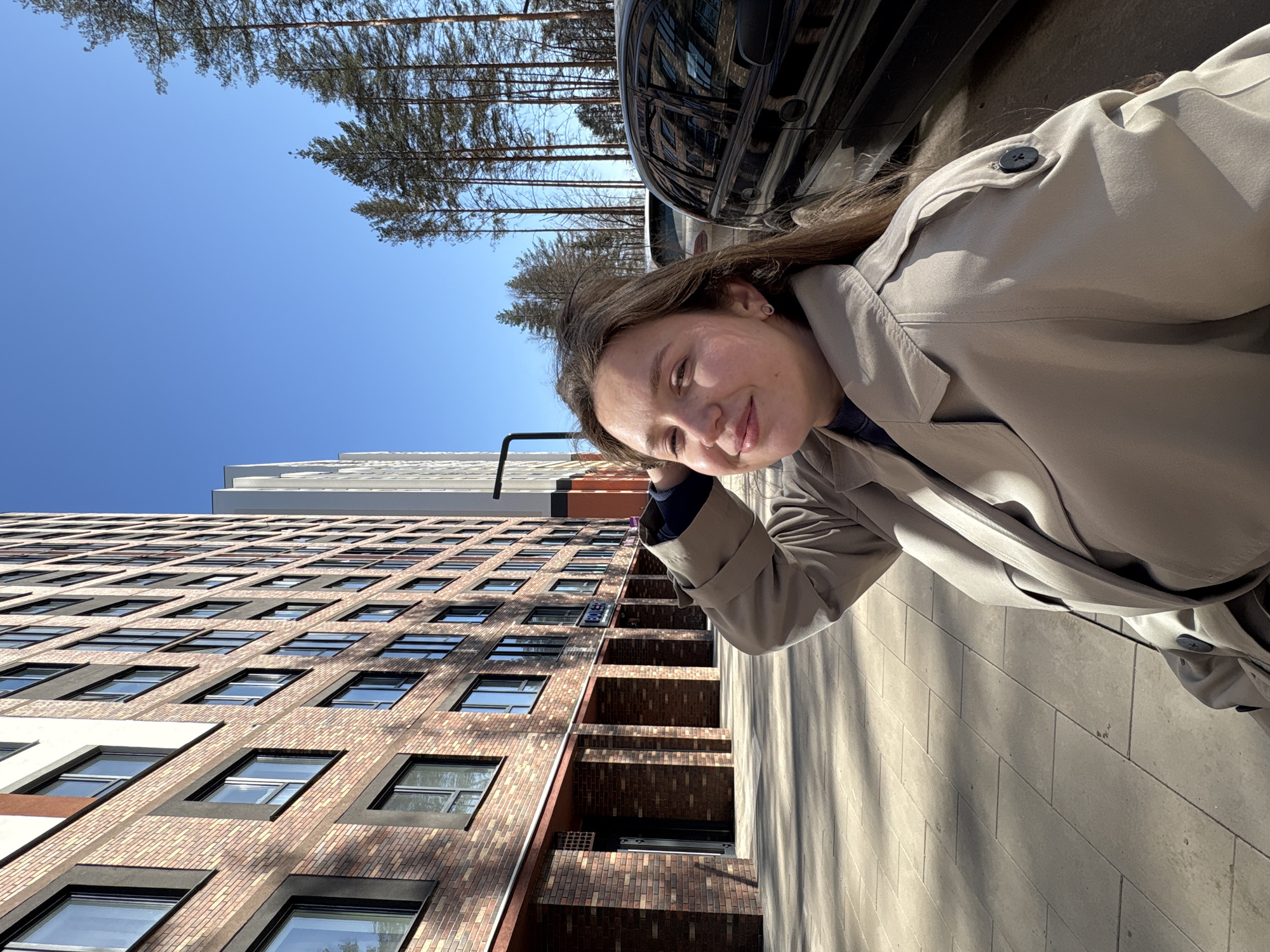 A woman in a beige trench coat stands on a paved sidewalk in Izhevsk, Russia. Behind her, a tall brick building with numerous windows rises against a clear blue sky. Bare trees stand in the background behind a black car.