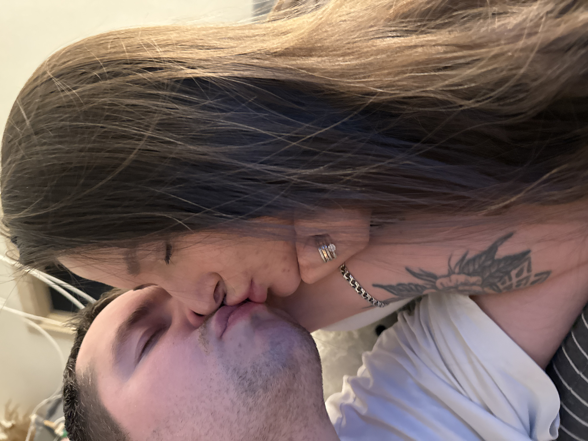 Two individuals, a man and a woman, are captured in an intimate moment, kissing passionately with their eyes closed. The image appears to be taken indoors, likely in a casual home setting in Akulovo (Gp Kubinka), Russia.
The woman is positioned above the man, leaning in for the kiss. She has long, dark brown hair with lighter, perhaps blonde, highlights visible, flowing across the upper and right portions of the frame. On her left ring finger, she wears a ring with multiple visible stones. A silver chain necklace adorns her neck, and a prominent dark tattoo, featuring a floral or nature-inspired design with abstract elements, is visible on her right shoulder/neck area. She is wearing a light-colored shirt, possibly white.
The man, lying beneath her, has short, dark hair and what appears to be light stubble or a short beard. He is wearing a white shirt. Both individuals' eyes are closed, emphasizing the depth of their connection and the emotional intensity of the moment. The background is softly blurred, showing light-colored walls or surfaces, reinforcing the indoor, personal atmosphere. There is no visible text in the image, and the lighting suggests indoor ambient light rather than specific time of day or outdoor conditions. The overall mood is one of affection, intimacy, and love.
