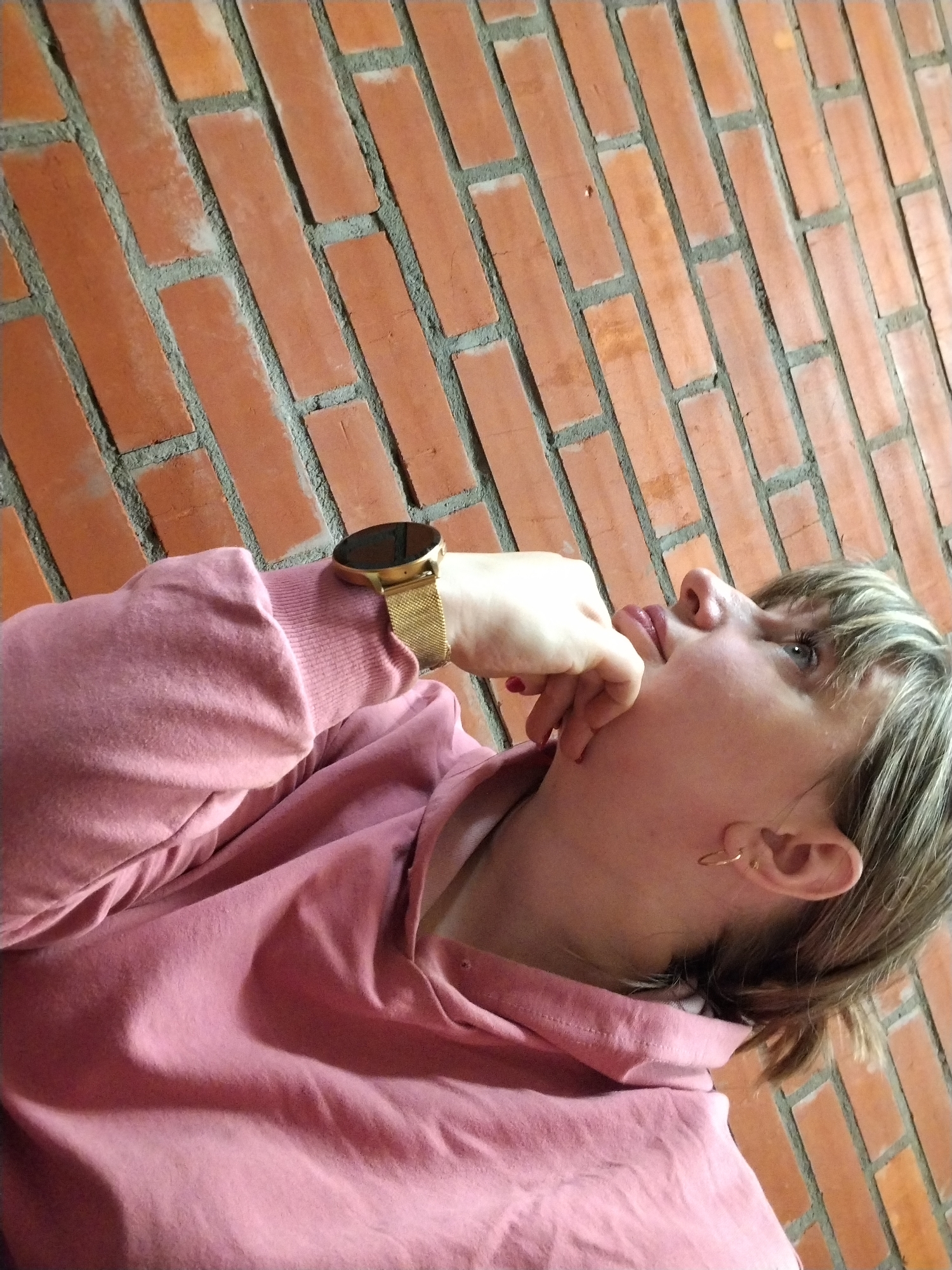 The image captures a person, likely female, in profile against a red brick wall. The person's face is turned upwards and to the right, with a thoughtful or pensive expression, and her gaze is directed out of frame. Her light-colored hair, possibly blonde or light brown, includes bangs across her forehead. She is wearing a soft pink hooded sweatshirt, which covers her body and arms. Her right hand is raised to her face, with fingers resting gently near her mouth and chin, reinforcing a contemplative mood.
On her left wrist, she wears a gold-colored smartwatch with a distinctive mesh band. A small gold hoop earring is visible in her right ear. The background consists of a uniformly patterned red brick wall with grey mortar, appearing at an angle due to the shot composition. The lighting is even, suggesting either an indoor setting or diffuse natural light. There is no visible text in the image. The general impression is one of quiet reflection.