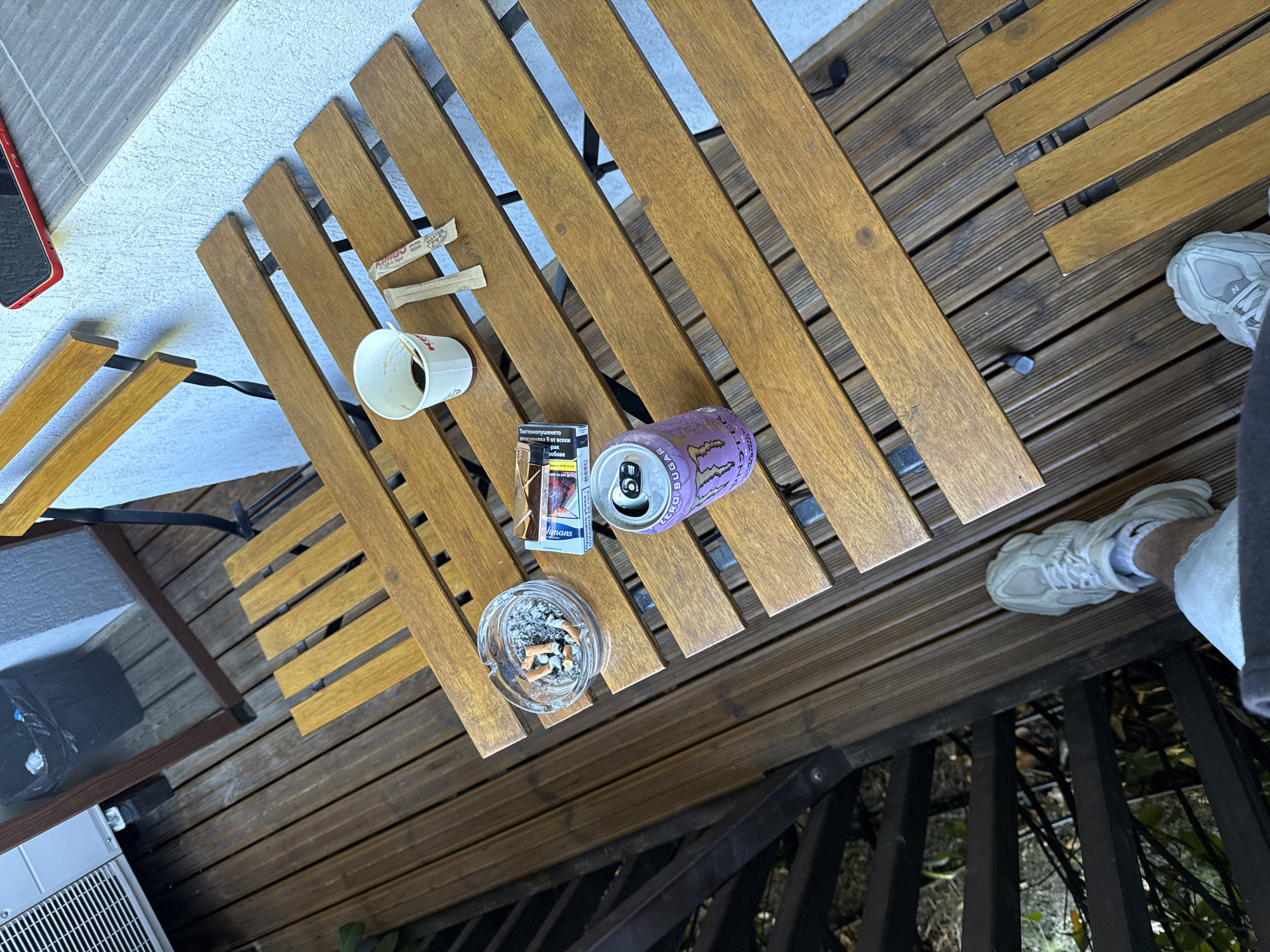 The image presents an elevated, oblique view of a scene set on a wooden deck or patio, with elements suggesting a casual, lived-in environment.
**Objects:**
* A rectangular, slatted wooden table is centrally positioned, bearing an assortment of items.
* These items include a half-empty paper coffee cup, two sugar packets, a pack of cigarettes, an ashtray filled with cigarette butts, and an open can of Monster Energy drink.
* A red cell phone rests near the edge of the table.
* A partial view of a white air conditioning unit is visible in the lower left corner.
**People:**
* Only the feet and lower legs of a person wearing white sneakers and shorts are visible in the lower right corner.
**Environment:**
* The setting appears to be an outdoor area with wooden decking.
* Black metal railings are visible in the background, with glimpses of greenery beyond.
* The location, based on the cigarette pack, is Veliko Tarnovo, Bulgaria.
**Inferences:**
* The scene suggests a relaxed setting, possibly a balcony or patio.
* The presence of cigarettes, coffee, and an energy drink implies activities involving smoking, caffeine consumption, and possibly a need for energy.
* The partial view of a person suggests that someone is present and interacting with the environment.