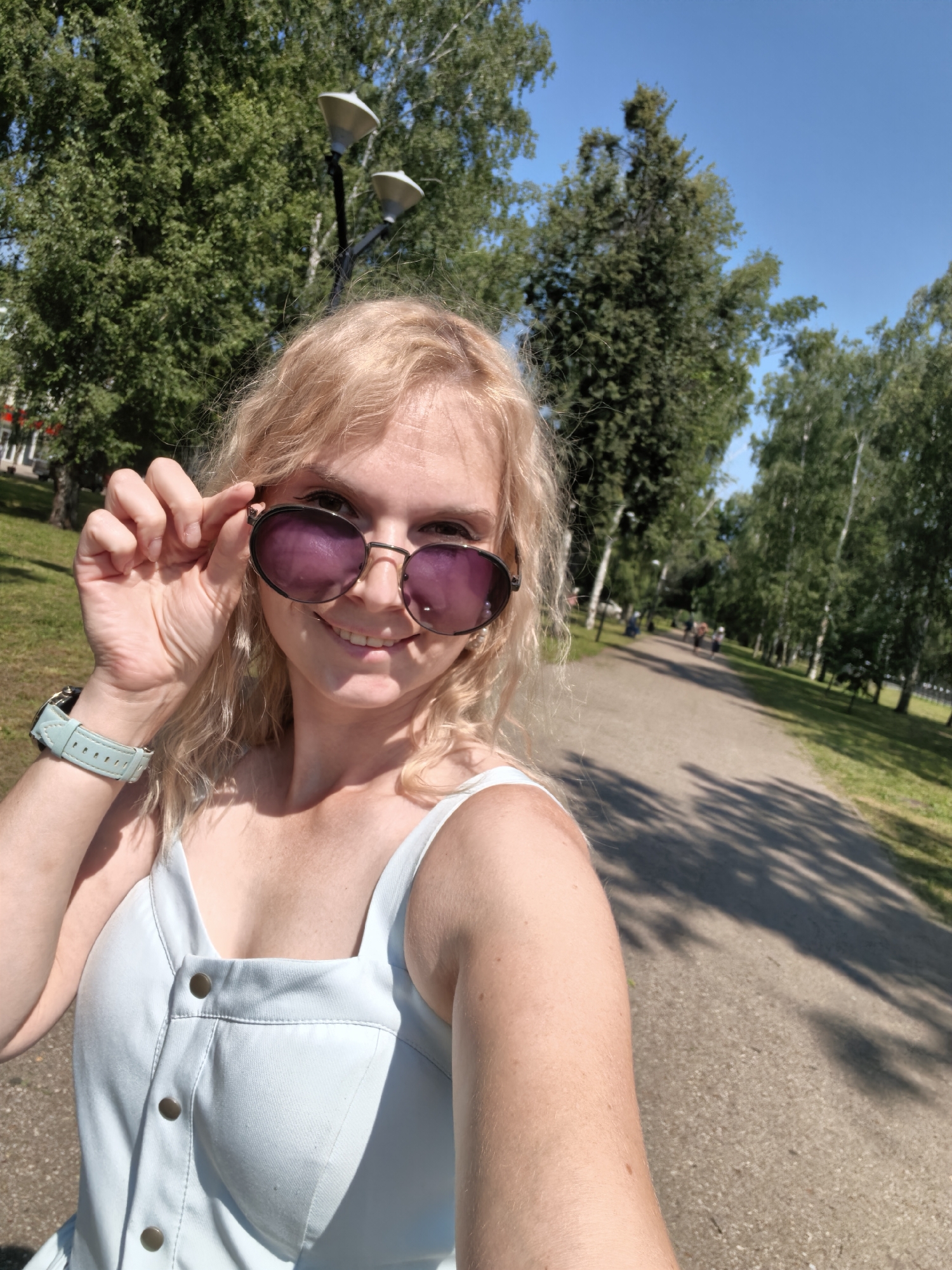 A young woman with blonde, wavy hair is taking a selfie in an outdoor park. She is wearing a light blue, button-down dress and sunglasses with purple lenses. She is holding the edge of her sunglasses with her right hand. A light blue watch is visible on her left wrist. The background features a paved walkway lined with trees and green grass, indicating a park setting. The sky is clear and blue, suggesting a sunny day. The location is identified as Sterlitamak, Russia.