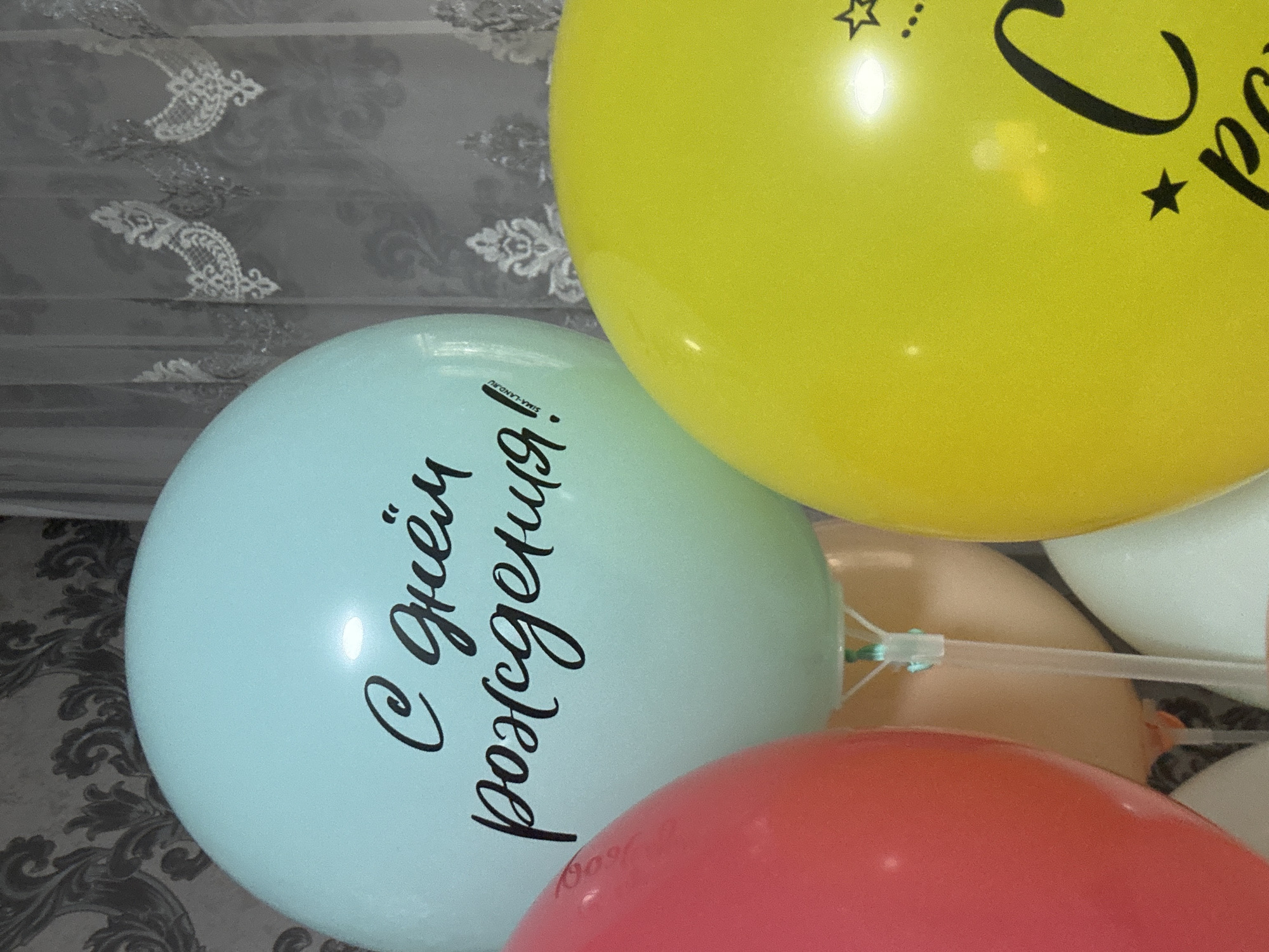 This is a close-up, eye-level, indoor photograph of several colorful balloons, likely a birthday decoration. The main focus is on a large, mint green balloon with black script writing that reads "С днём рождения!" (Happy Birthday!). To the left of this balloon, a portion of a bright pink balloon is visible, and to the right, a large, bright yellow balloon with black stars and lettering that appears to be "С днём" is prominent. In the background, there are sheer curtains with a floral pattern and a darker, patterned wallpaper. The lighting is adequate, highlighting the balloons and their inscriptions. The image does not provide any information about a specific location in the city of Марковский, Russia, or any people.