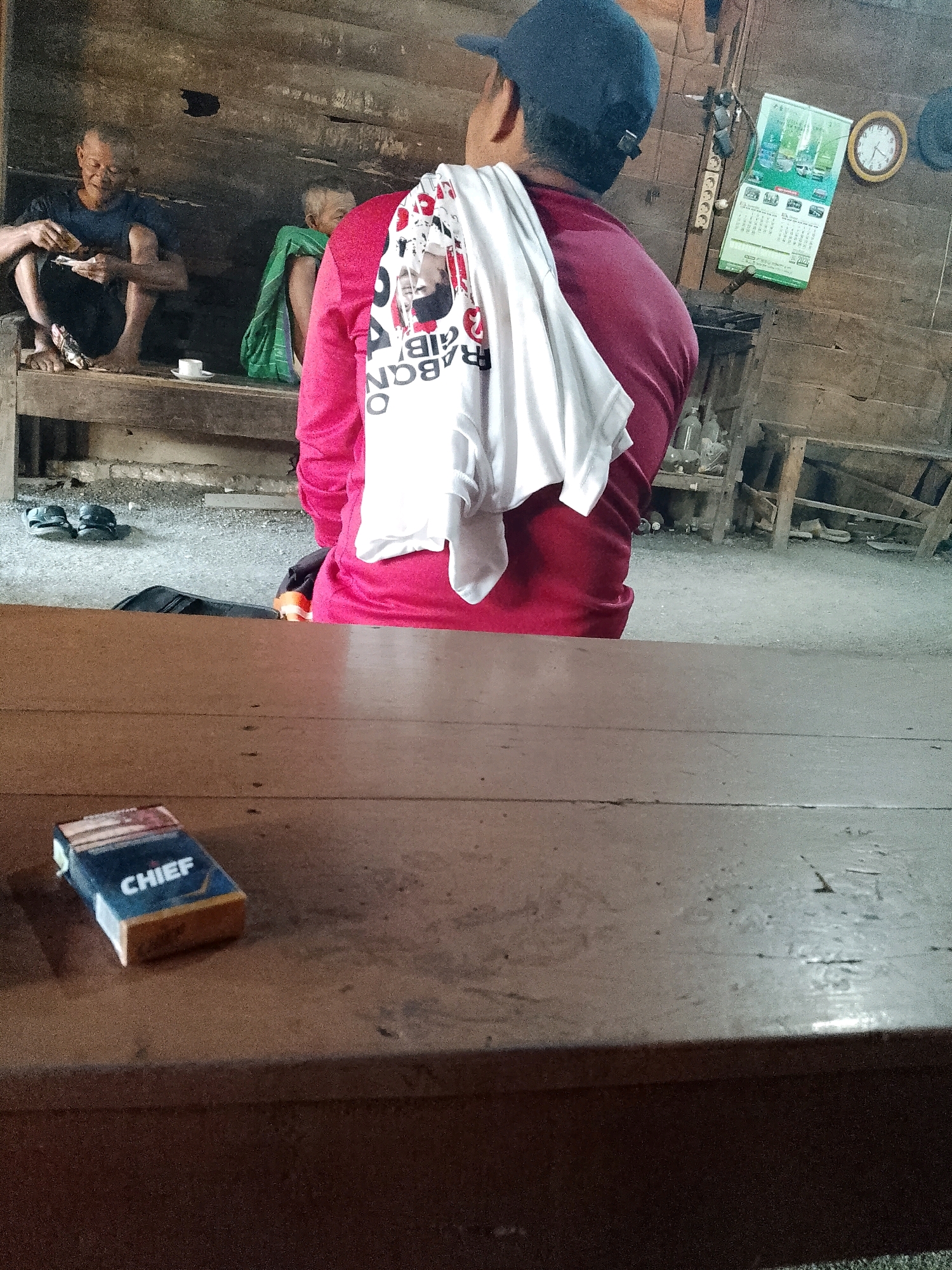 In Wadang, Indonesia, a man wearing a red long-sleeved shirt and a blue baseball cap is shown from the back, with a white t-shirt draped over his shoulders. The t-shirt has a graphic print. In the background, two older men are seated on a wooden platform. The man on the left, wearing a dark shirt, is holding something small in his hands. He is barefoot. Next to him, a small white cup sits on the platform. Another man, partially obscured by the first, is wearing a green cloth. Further back, a calendar hangs on a wooden wall, alongside an analog clock. Various wooden structures and shelves are visible in the background, along with some bottles and what appears to be debris on the floor. In the foreground, a pack of "CHIEF" cigarettes rests on a wooden table. Sandals are placed on the ground near the seated men. The scene appears to be an outdoor or semi-outdoor setting, likely a rural or less developed area, given the rustic construction materials and general environment.
