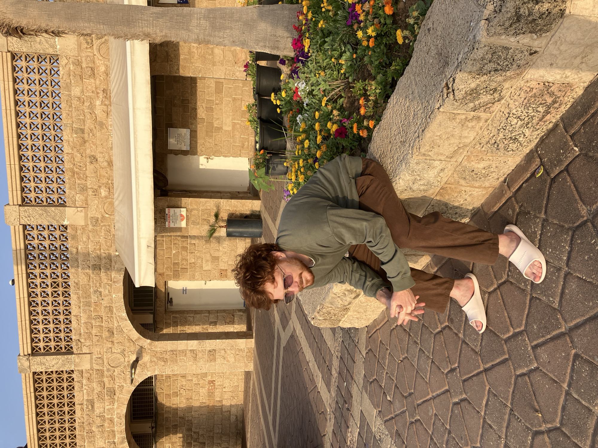 A man with light skin, auburn hair, a light beard, and sunglasses is sitting cross-legged on a stone bench in Tel Aviv, Israel. He is wearing a long-sleeved green shirt, brown pants, and white sandals. He is sitting adjacent to a flower bed filled with colorful flowers in black pots. Behind him is a building made of light brown stone bricks, featuring archways and decorative patterned windows. The ground is paved with interlocking brown and tan stones. The sun is shining, casting shadows in the scene.