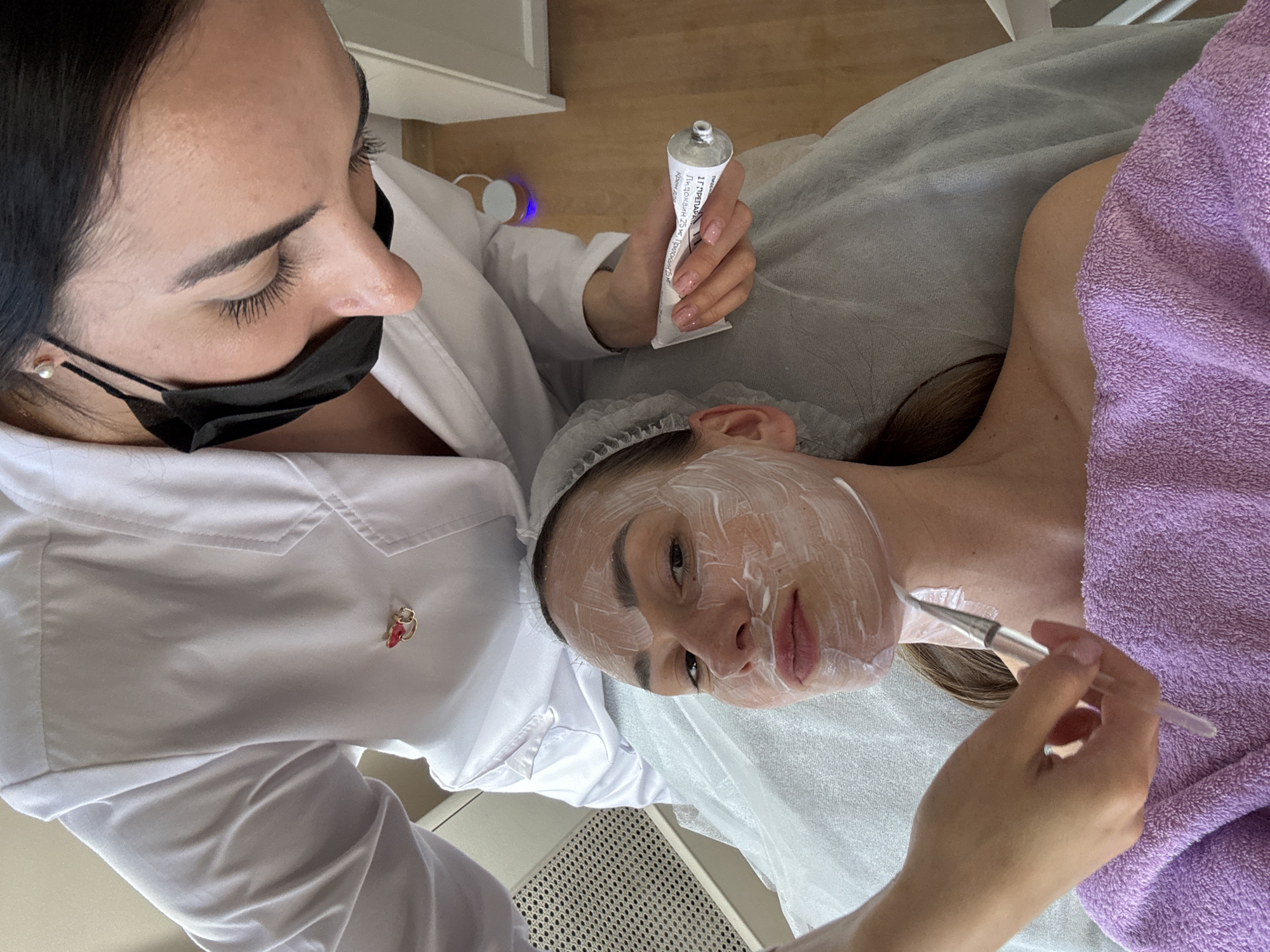 This close-up, high-angle shot captures a cosmetology procedure in what appears to be a salon or medical spa in Sochi, Russia. Two women are the primary subjects.
On the left, an aesthetician, wearing a white medical coat or uniform and a black face mask, looks down intently at her client. Her dark hair is pulled back, and she wears a small pearl earring in her right ear. A small, decorative brooch with red and gold elements is pinned to her uniform. Her right hand holds a white tube of product, while her left hand is out of frame, likely holding the brush. Her fingernails have a light pink, possibly glittery, polish.
The client, positioned horizontally, is receiving a facial treatment. Her face is largely covered with a white cream or mask, which has been applied with visible brush strokes. She wears a disposable hairnet to keep her hair away from her face, and her body is covered with a purple terry towel and a disposable white sheet or covering. Her eyes are open, looking slightly upwards towards the aesthetician. Her expression appears neutral or relaxed. Her left arm is visible, and her hand holds a brush, seemingly applying more product to her own neck area, or perhaps the aesthetician is guiding her hand.
In the background, a small device with a blue light is faintly visible. The floor appears to be light-colored wood.
Visible text on the product tube held by the aesthetician is in Cyrillic. It reads "ГИДРОГЕЛЬ" (Hydrogel), "ПАНАВИР 25 г" (Panavir 25 g), and "произведено в России" (produced in Russia). Panavir is often associated with antiviral treatments, available in gel form.
The scene suggests a professional skincare treatment, likely taking place during the daytime in a well-lit indoor environment.