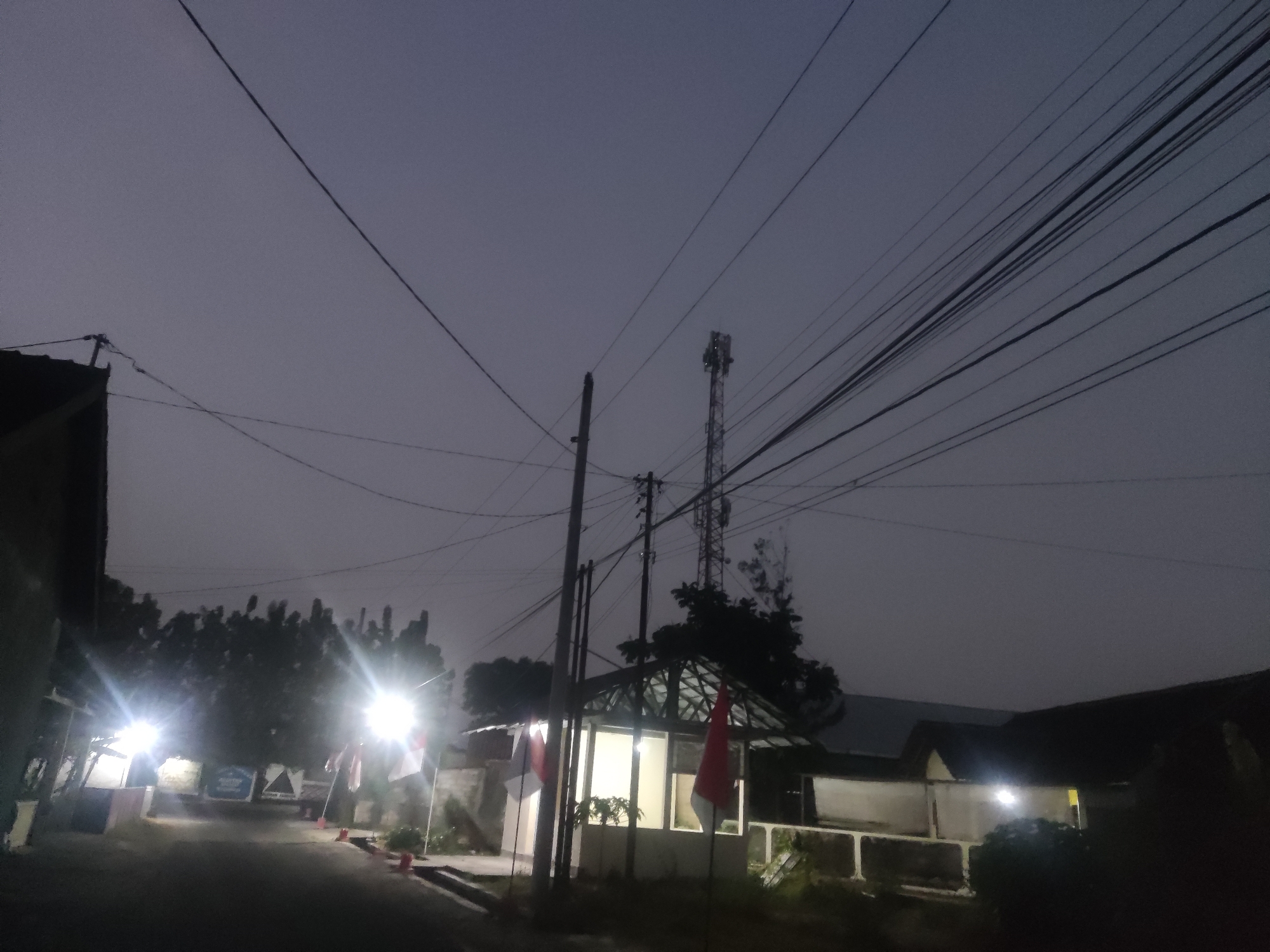 The image depicts a twilight scene on a street in Trihanggo, Indonesia, with several buildings and a communication tower visible. The sky is a muted gray, suggesting either dawn or dusk. A road curves through the foreground, flanked by low-rise structures. On the left, a building with a dark roof and a portion of a light-colored wall is visible, with bright lights emanating from within or near it. Further down the street, two red and white flags, likely the Indonesian national flag, are displayed on flagpoles in front of a building with a covered outdoor area. This structure has windows that are illuminated from within. To the right of this building, another structure with a dark roof is partially visible, also with interior lights. A prominent communication tower stands in the background, silhouetted against the sky. Numerous power lines crisscross the frame, adding to the urban or semi-urban setting. Trees are present in the mid-ground, appearing dark against the twilight sky. The overall impression is one of a quiet, early morning or late evening in a residential or mixed-use area.