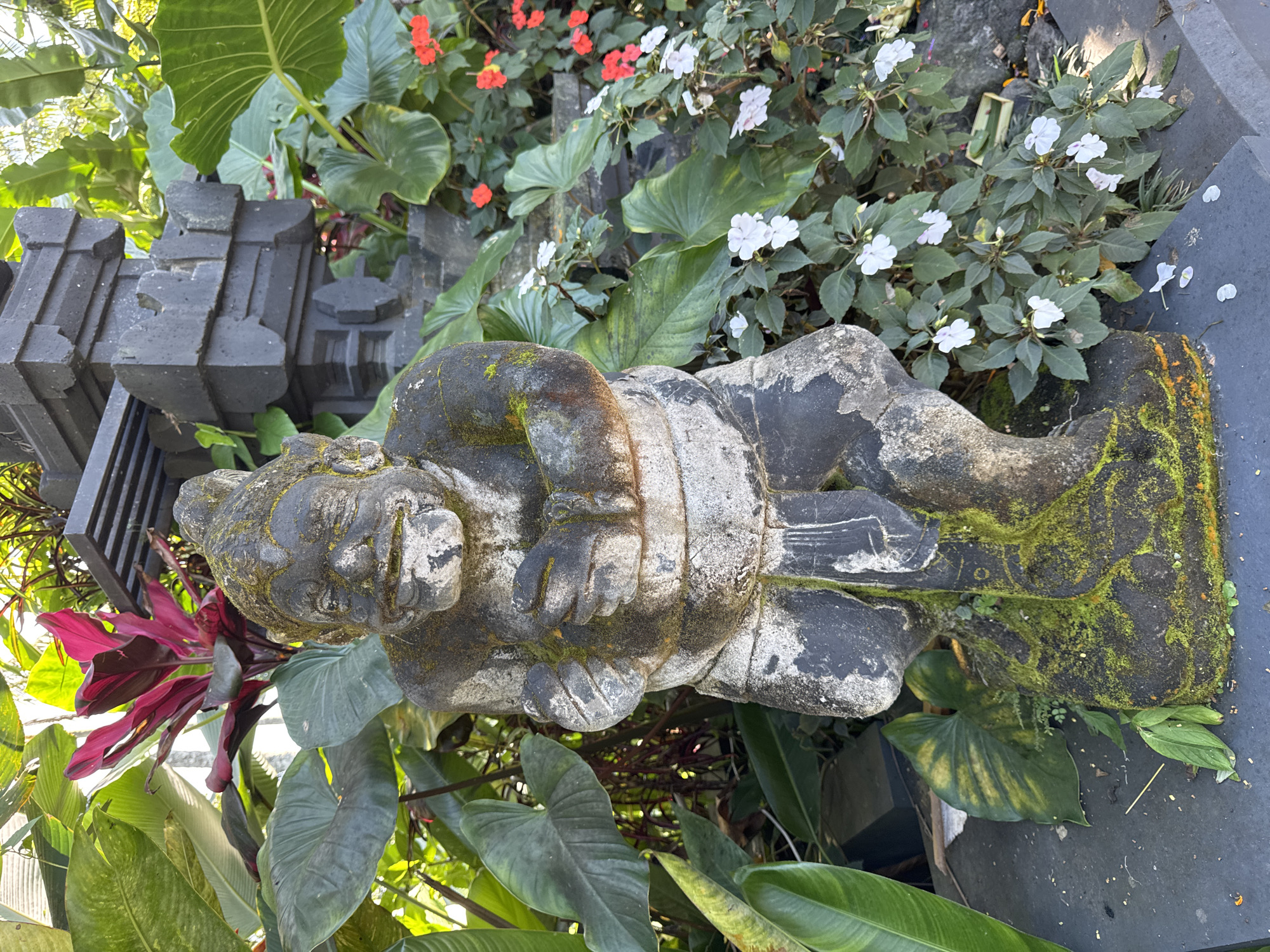 The image shows a weathered stone statue of a squatting figure, covered with moss, set amidst lush vegetation. The statue is positioned near a dark, multi-tiered stone structure resembling a gate or temple facade. The surroundings are rich with varied plant life, including large-leafed plants, red and white flowering plants. This scene is likely located in Rendang, Indonesia.