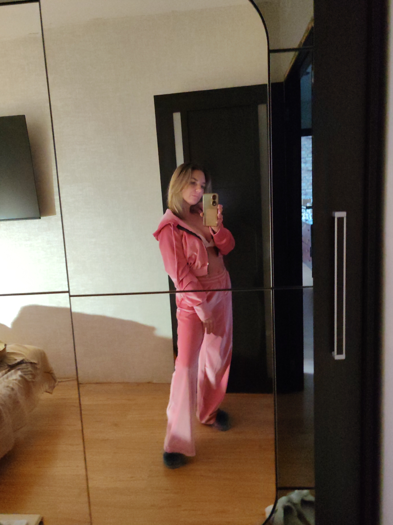 A young woman wearing a pink velour tracksuit is taking a selfie in a mirror. The mirror is part of a wardrobe located in a room with a wooden floor, a light-colored textured wall, a bed with tan bedding, and a black doorway. The location is likely indoors. The photo does not include enough information to determine if it is located in the city of Petropavlovsk-Kamchatsky, Russia.
