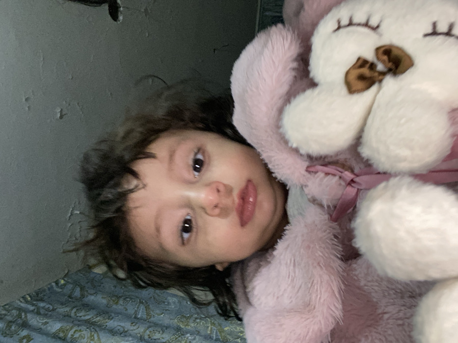 A young child with brown, slightly messy hair lies on a patterned surface, possibly a bed. The child gazes directly at the camera with big, brown eyes. She's wearing a pink, fuzzy garment and is cuddling a large, white stuffed flower with a brown center and a pink ribbon. The background features a textured gray wall, adding to the cozy, indoor setting.
