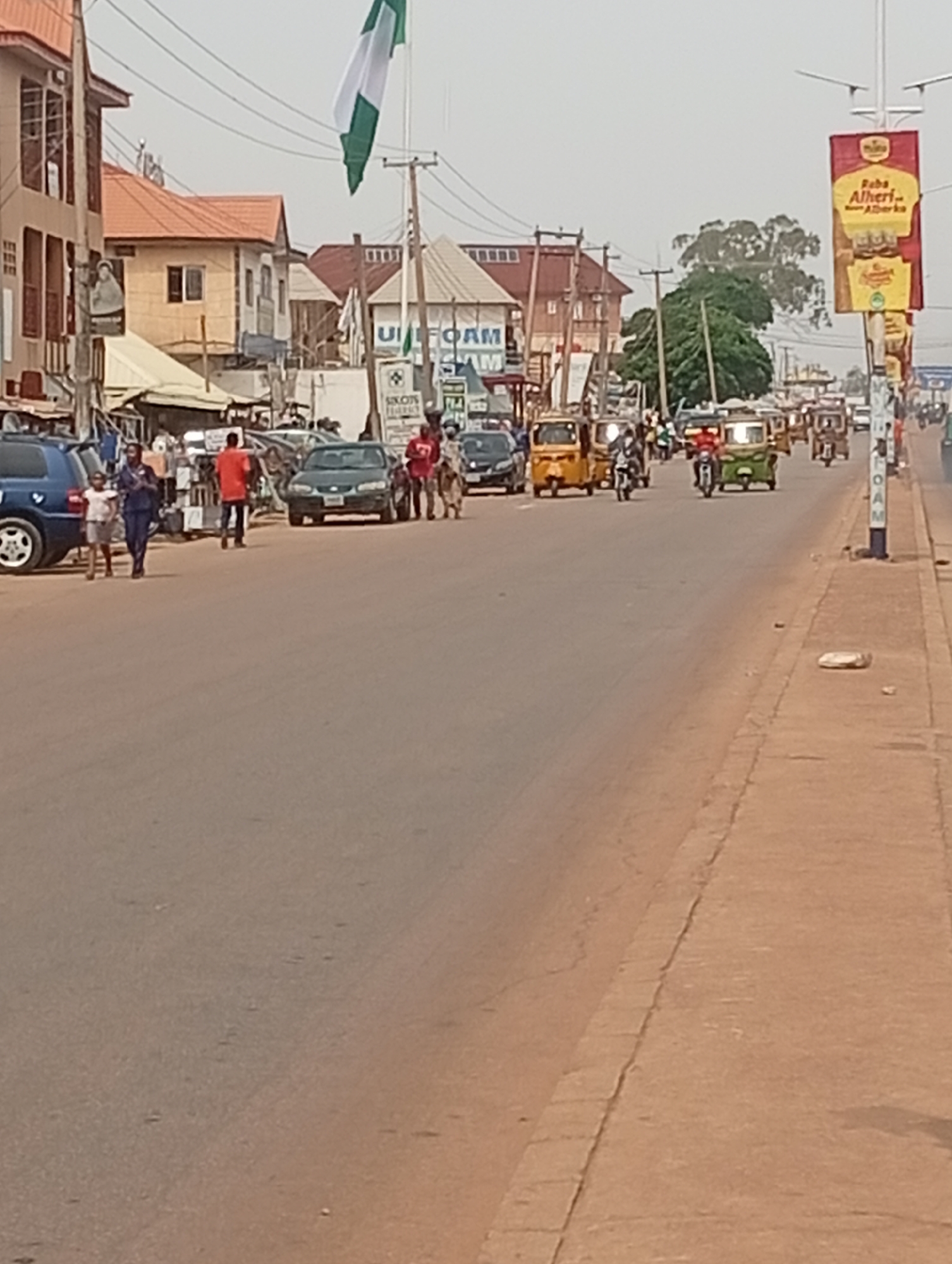 The image depicts a street scene in Sabon Tasha, Nigeria. A wide asphalt road dominates the foreground, extending into the distance, slightly blurred by the heat or atmospheric conditions.
On the left side of the road, a blue SUV is parked near the sidewalk. Several people are walking along the left side of the street, including an adult in a dark outfit holding hands with a child in a white top. Another adult in an orange shirt is seen walking further down the street.
Various vehicles, including cars, yellow and green motorized rickshaws, and motorcycles, are moving along the road. Buildings with orange roofs are visible along the left side of the street. A large Nigerian flag is prominently displayed. A billboard with the text "Baba Alheri... Abarka" is positioned on the right side of the street near power lines. Further down the street, a sign that reads "UI Foam" is visible.