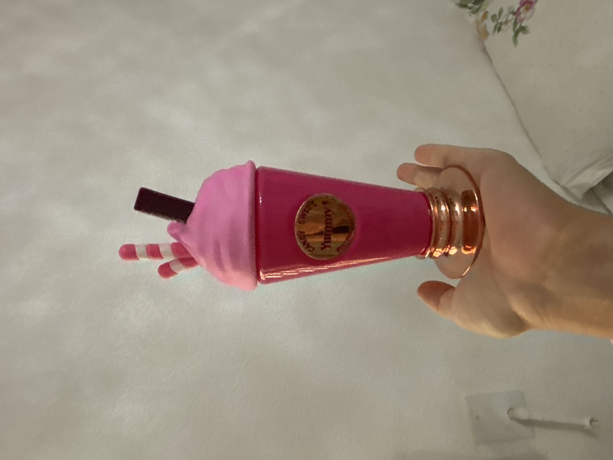 A hand holds a bright pink, cup-shaped object resembling a milkshake or dessert. The object has a swirled pink top, two pink and white striped candy-like sticks, and a dark brown rectangular piece on top, possibly representing a chocolate bar. A gold circular label with the word "Yummy" in gold lettering is affixed to the front of the cup. The cup is supported by a metallic gold base. The background is a plain, textured light-colored wall with a white electrical outlet visible on the left side. The image appears to be taken indoors, possibly in a residential setting in Orihuela, Spain, given the context.