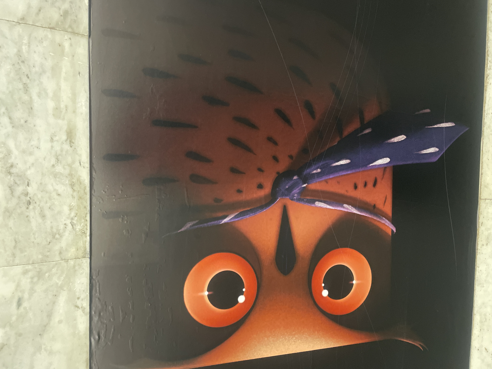The image shows a close-up of an owl illustration with prominent orange eyes and a purple bandana. The background is dark brown and black. The illustration appears to be on a flat, possibly rectangular surface. No evidence suggests that the image is in Yerevan, Armenia.