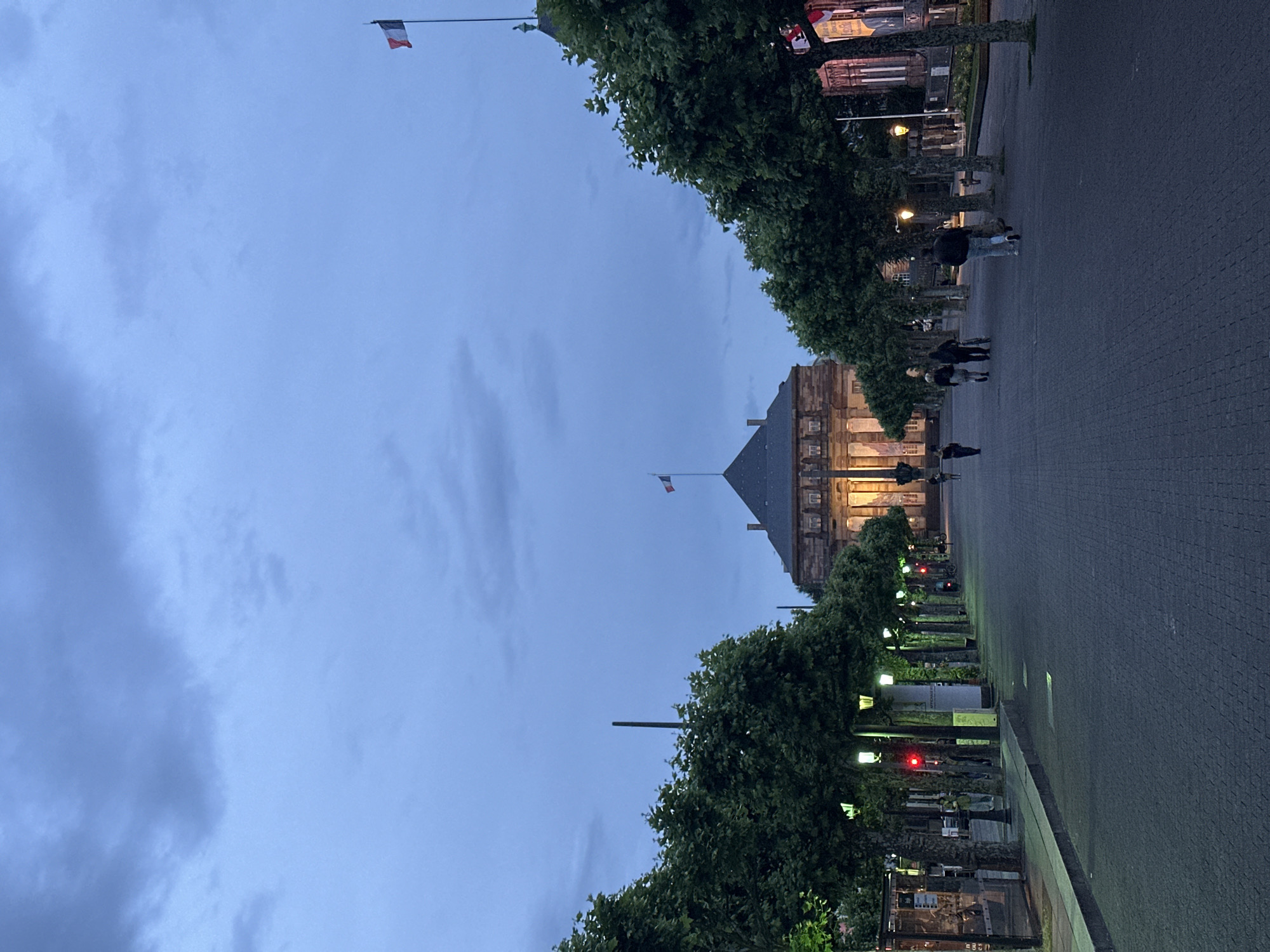 An evening view captures a wide, tree-lined avenue or plaza in Strasbourg, France, at dusk. The sky is a soft, cloudy blue-grey, indicating the transition between day and night. Artificial lights illuminate a grand, historic building in the background, featuring multiple windows and a distinctive dark roof, suggestive of European architecture.
Along both sides of the paved pathway, rows of lush green trees frame the scene. Scattered streetlights and other light fixtures are visible among the foliage, some emitting a warm glow, while two distinct red lights appear on the right side, possibly traffic signals or decorative elements.
Several blurred figures of people are seen walking casually on the dark, paved ground, suggesting a peaceful, everyday activity. Two French flags (blue, white, red) are prominent against the sky: one larger flag on a flagpole in the upper left, and a smaller one flying from the historic building in the distance. The overall atmosphere is quiet and tranquil. No discernible text is visible.
