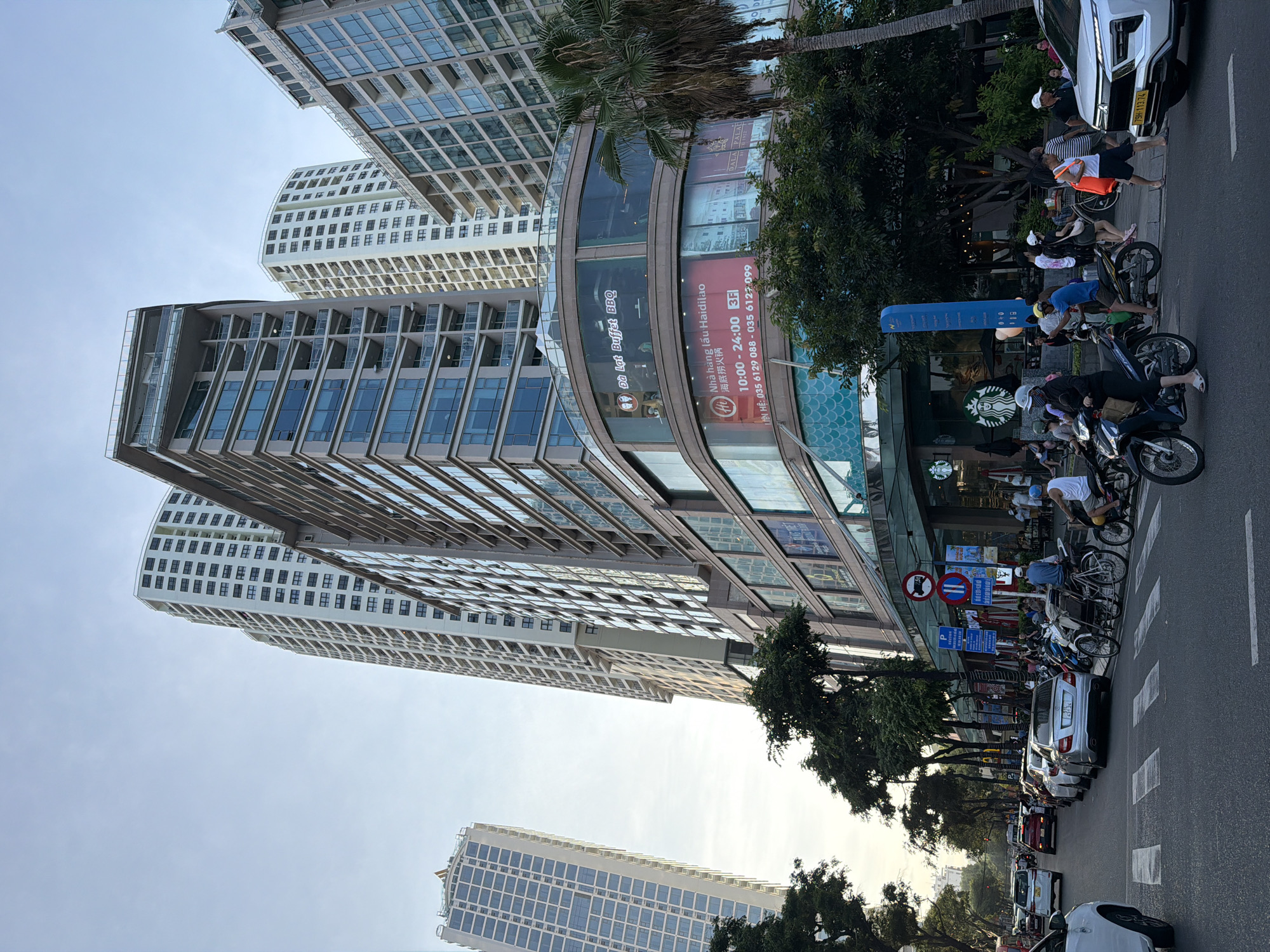 This image depicts a bustling urban street scene in Nha Trang Ward, Vietnam, specifically showing the Gold Coast building at 01 Trần Hưng Đạo. The central focus is a multi-story commercial and residential complex featuring a curved glass facade on its lower levels. A sign for "Đà Lạt Buffet BBQ" is visible on the second floor, and below it, "Nhà hàng lẩu Haidilao" is advertised on the third floor with operating hours (10:00 - 24:00) and two phone numbers (035 6129 088 and 035 6127 099). At street level, the green circular logo of a Starbucks coffee shop is prominent.
Behind this central building and to its sides, several tall, modern skyscrapers are visible. These buildings exhibit repetitive patterns of windows and balconies, indicative of high-rise residential or mixed-use developments. One tall, light-colored building with numerous dark-framed windows stands directly behind the central structure, while another, partially seen on the far left, also features a grid-like window design. A third tall building with glass balconies is present on the right edge of the frame.
The street in the foreground is an asphalt road with a marked white zebra crossing. Vehicular traffic includes numerous cars and motorcycles. A white Mercedes-Benz sedan (license plate 51L-431.24) is parked on the right side of the street. Further right, a white SUV (license plate 79H-113.74) is partially visible. Other cars, including red and white sedans, are present further down the road. Motorcycles are ubiquitous; several are parked along the sidewalk, and individuals are riding them, such as a person in a white helmet and mask on a black scooter on the left, and a man in a white tank top and blue helmet on a dark motorcycle on the right.
Pedestrians populate the sidewalks and are crossing the street. On the right, a man in a white tank top carrying an orange bag crosses the street barefoot. Another man in a blue shirt and white cap stands near a traditional bicycle taxi (xích lô) on the sidewalk. People are also gathered near the Starbucks entrance, some seated at outdoor tables. Green trees line the sidewalk, providing shade, and a tall palm tree is visible on the far right. Street signs, including a blue parking sign and a red no-stopping sign, are positioned on the sidewalk. The sky is a light, hazy blue, suggesting either late afternoon or early evening lighting conditions.