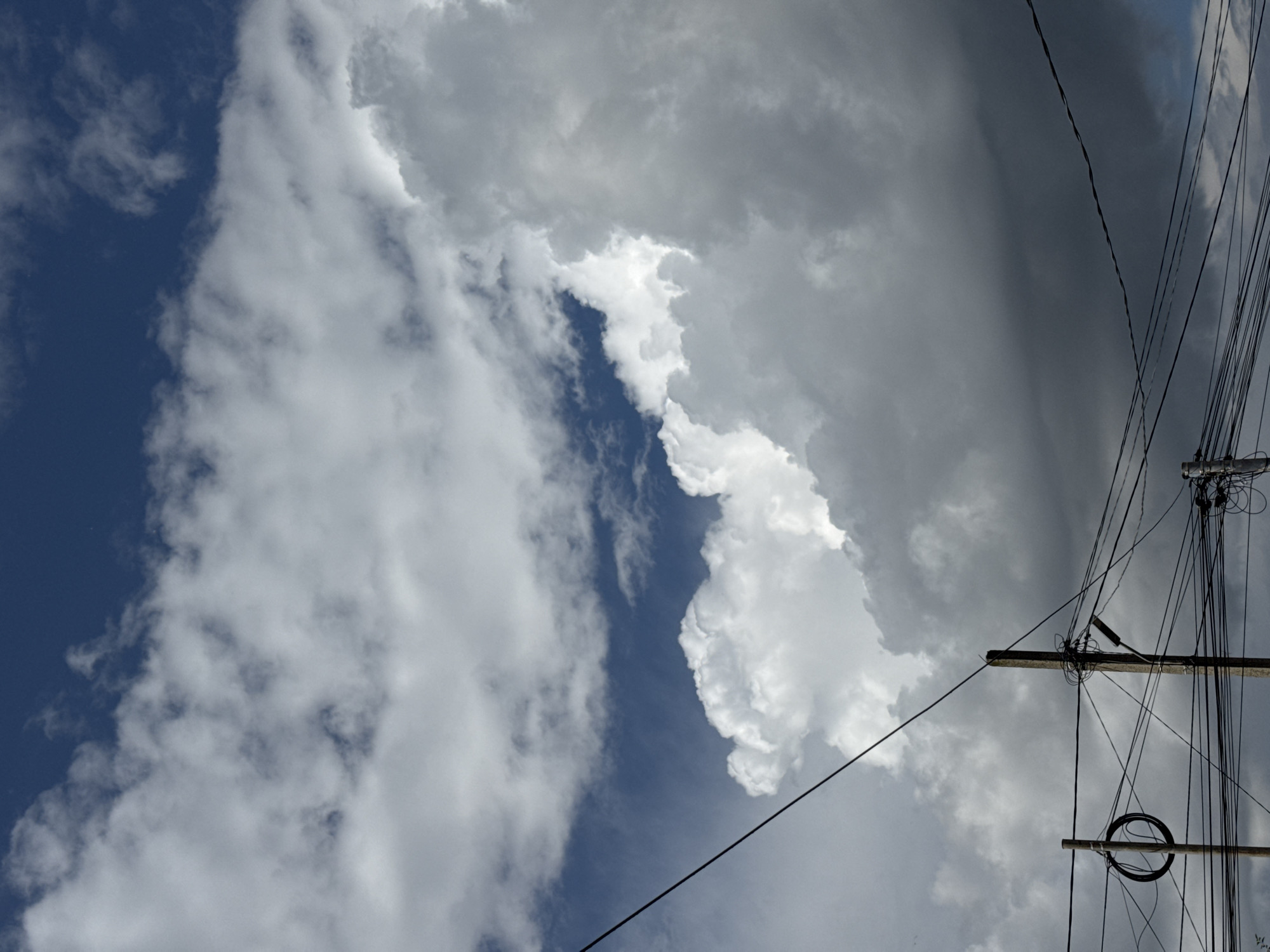 The image depicts a sky filled with dramatic clouds over a cityscape. The clouds are a mix of bright white and dark gray, suggesting a dynamic weather system. The sky transitions from a deep blue at the top to overcast gray towards the bottom. In the foreground, there are utility poles and a dense network of electrical wires crisscrossing the frame. One pole on the left has a circular object resembling a coil of cable attached to it. The scene appears to be an outdoor environment, possibly in a developed area with infrastructure for power distribution. The presence of clouds and overhead wires are the dominant visual elements. The image does not contain any discernible people or specific architectural features to pinpoint an exact location beyond a general urban or suburban setting.