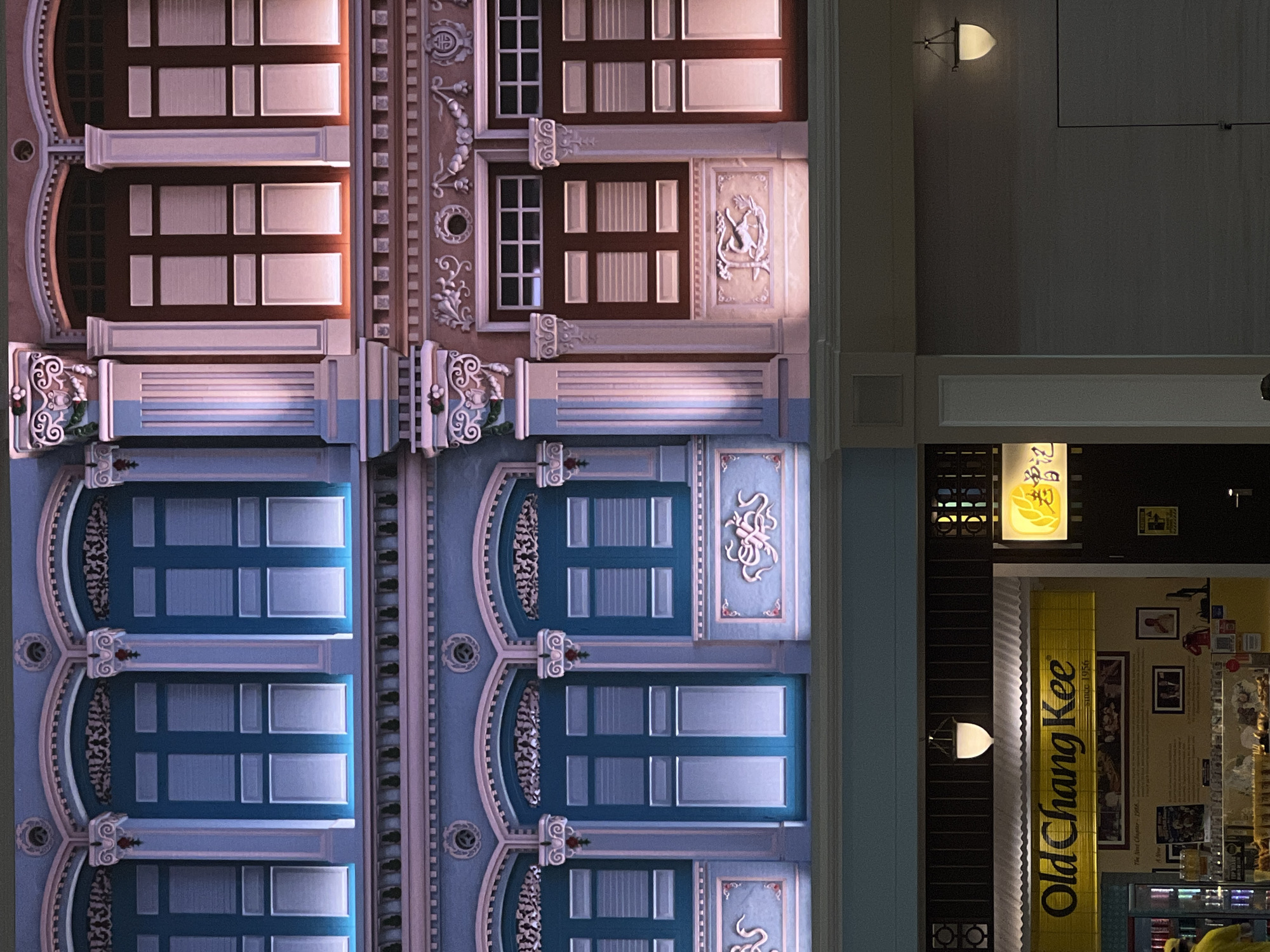 The image shows the exterior of the Old Chang Kee restaurant in Singapore, located at 178 Old Airport Road Food Centre. The facade features traditional shophouse architecture with ornate details and blue and brown shutters. The lighting suggests it is evening or night, with warm lights illuminating the building and a yellow sign for Old Chang Kee prominently displayed. The restaurant appears to be open for business, with patrons potentially visible inside or in the process of entering or exiting. The overall scene depicts a well-preserved historical building utilized as a commercial establishment, reflecting Singapore's rich cultural heritage.